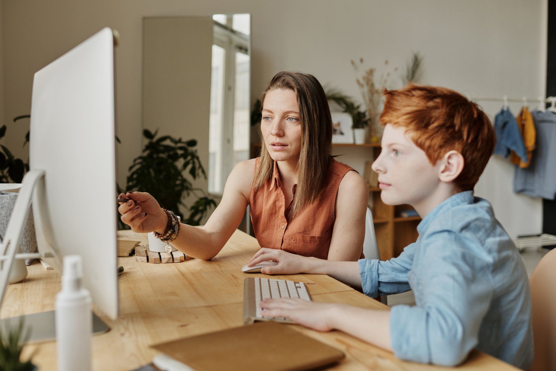 White woman and redheaded boy work at computer together. She uses pen to point to screen