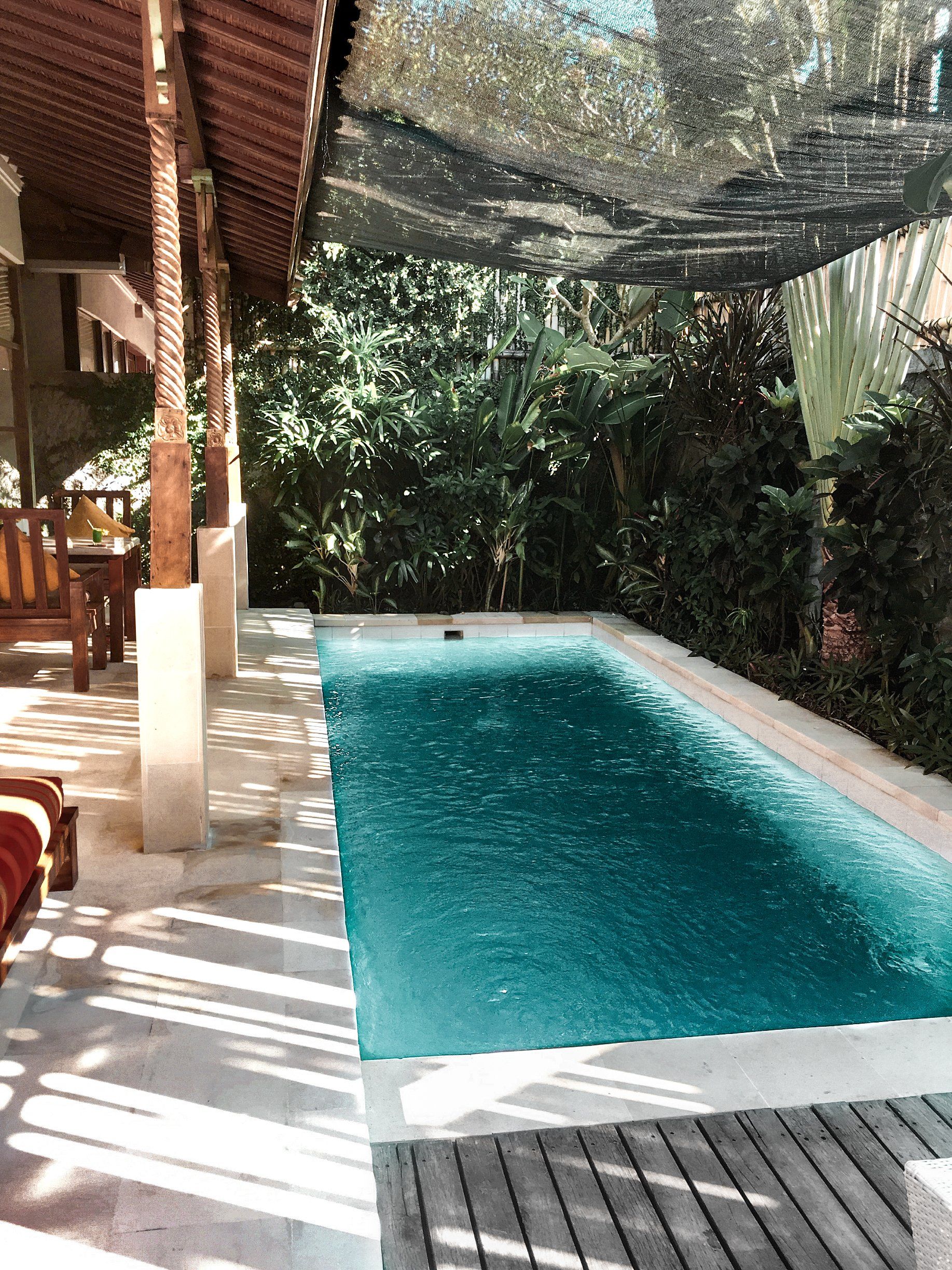 Pool under shaded patio, lush greenery backdrop, turquoise water, light wood deck.