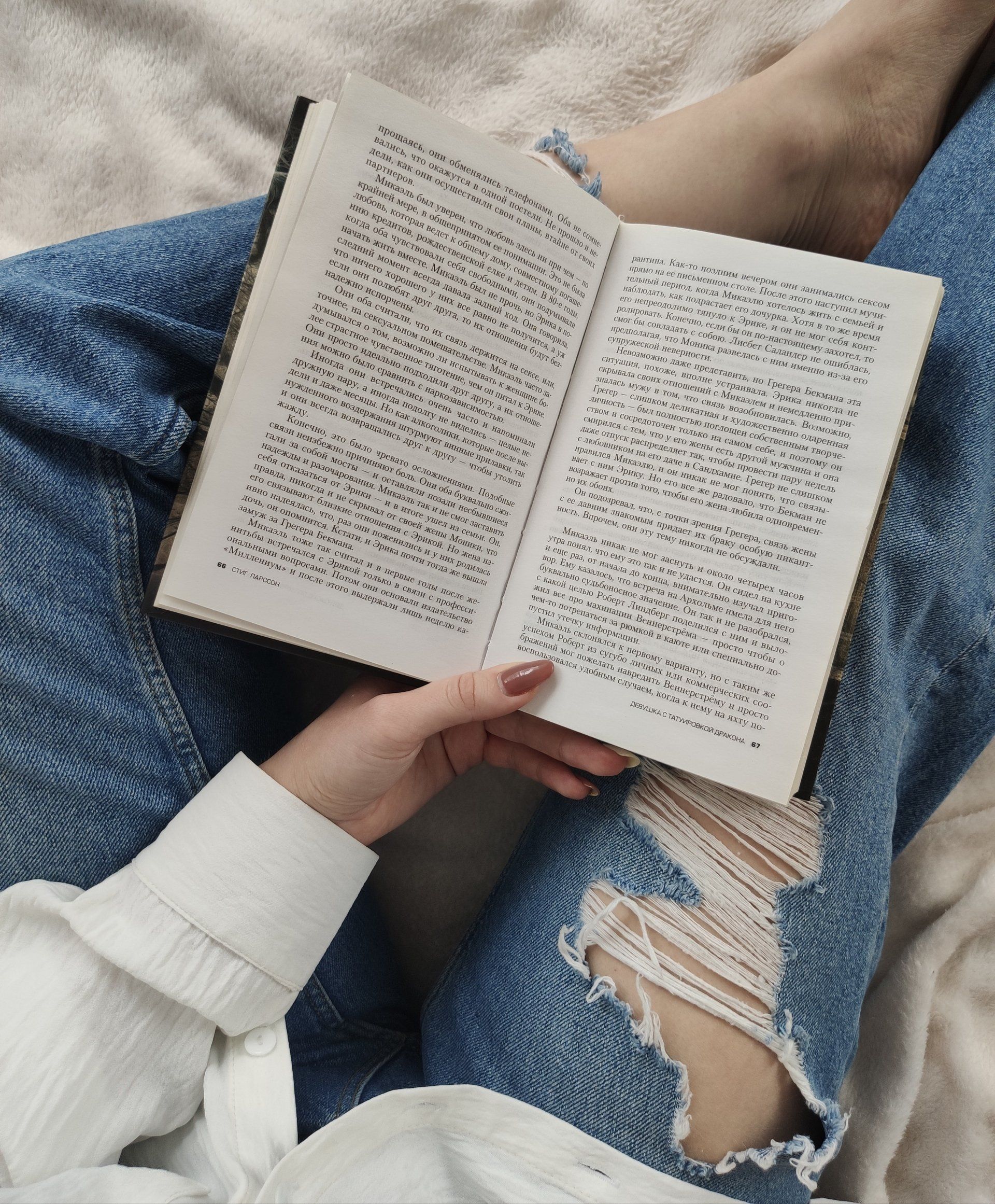 Una mujer con jeans rotos está leyendo un libro en una cama.