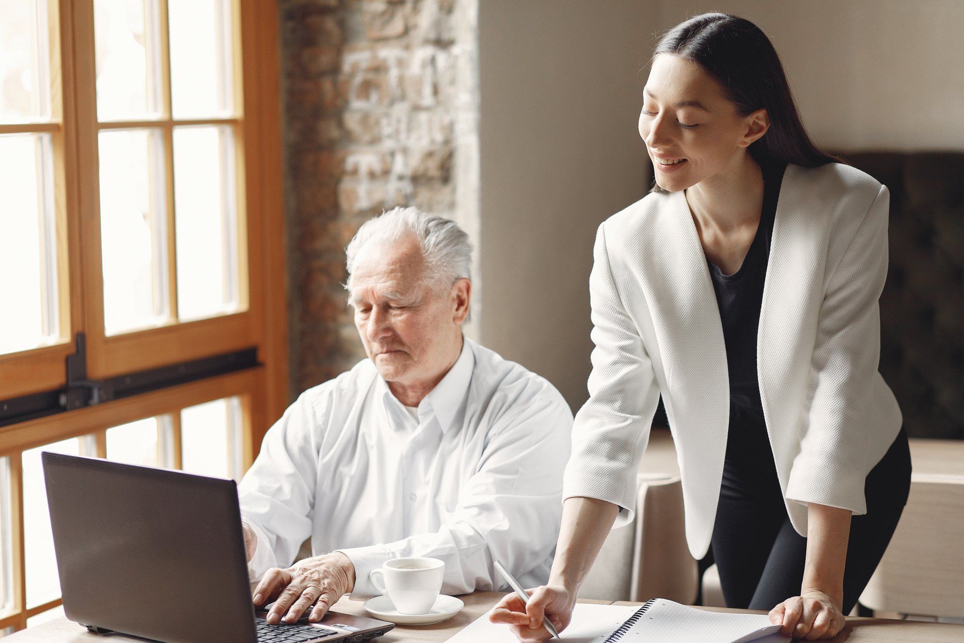A woman is standing next to an older man using a laptop computer.