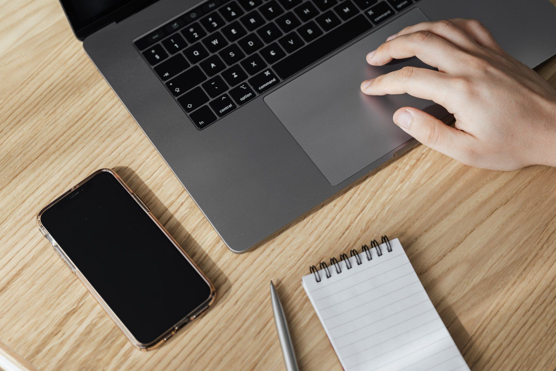 A person is typing on a laptop next to a cell phone and a notebook.