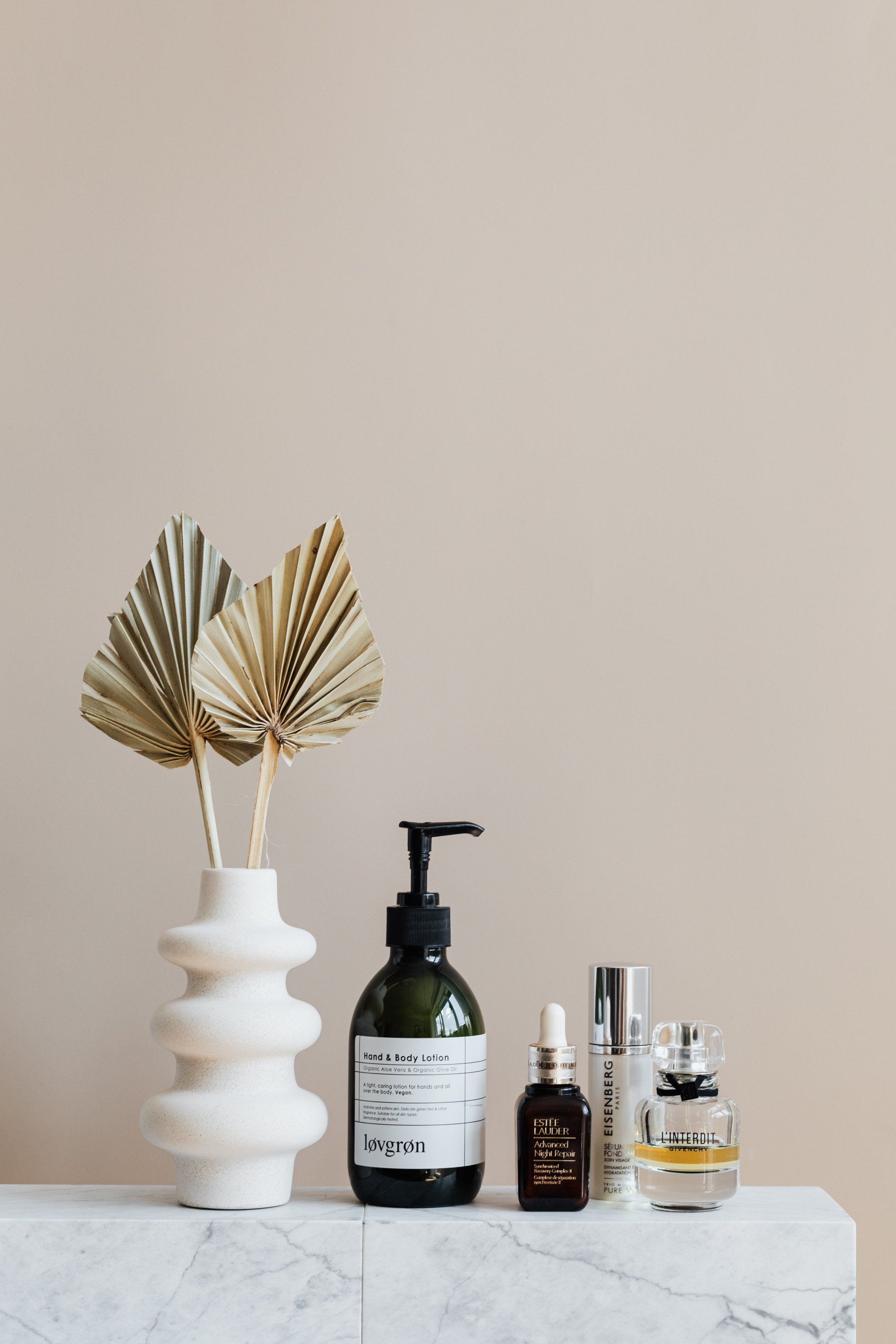 Skincare products and vase with dried palm leaves on a marble shelf against a beige wall.