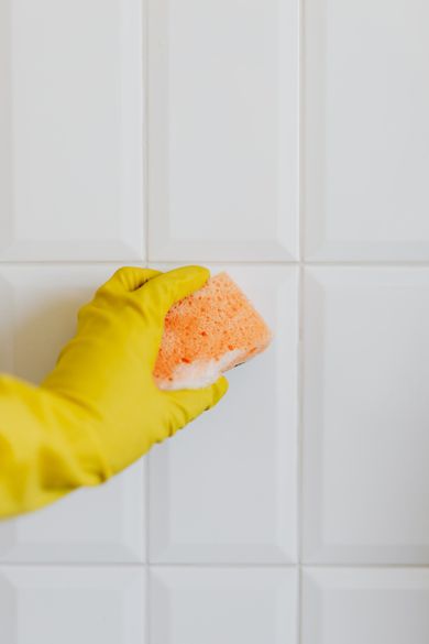 A person wearing yellow gloves is cleaning a white tile wall with a sponge.