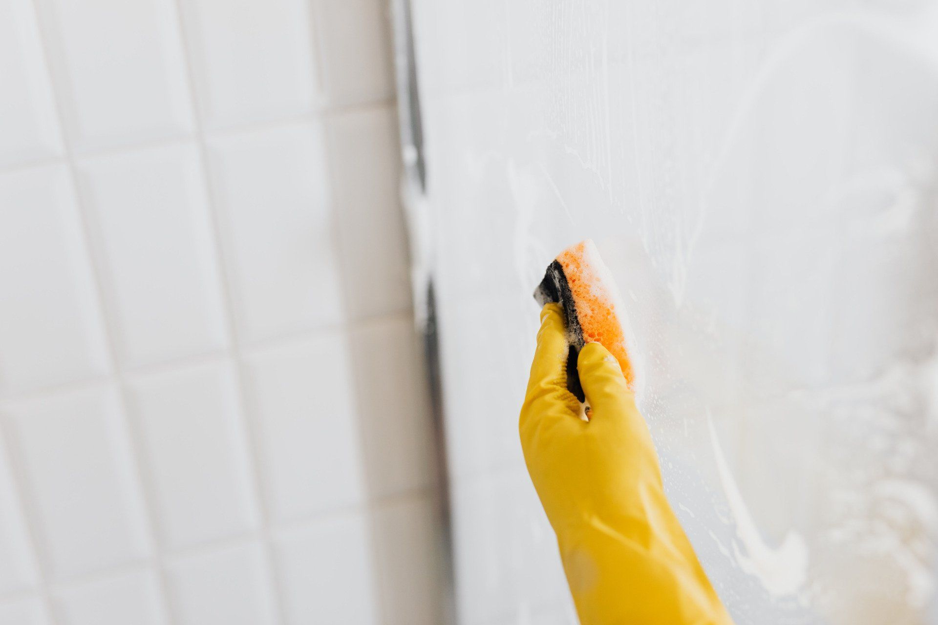 A person wearing a yellow glove is cleaning a shower door with a sponge.