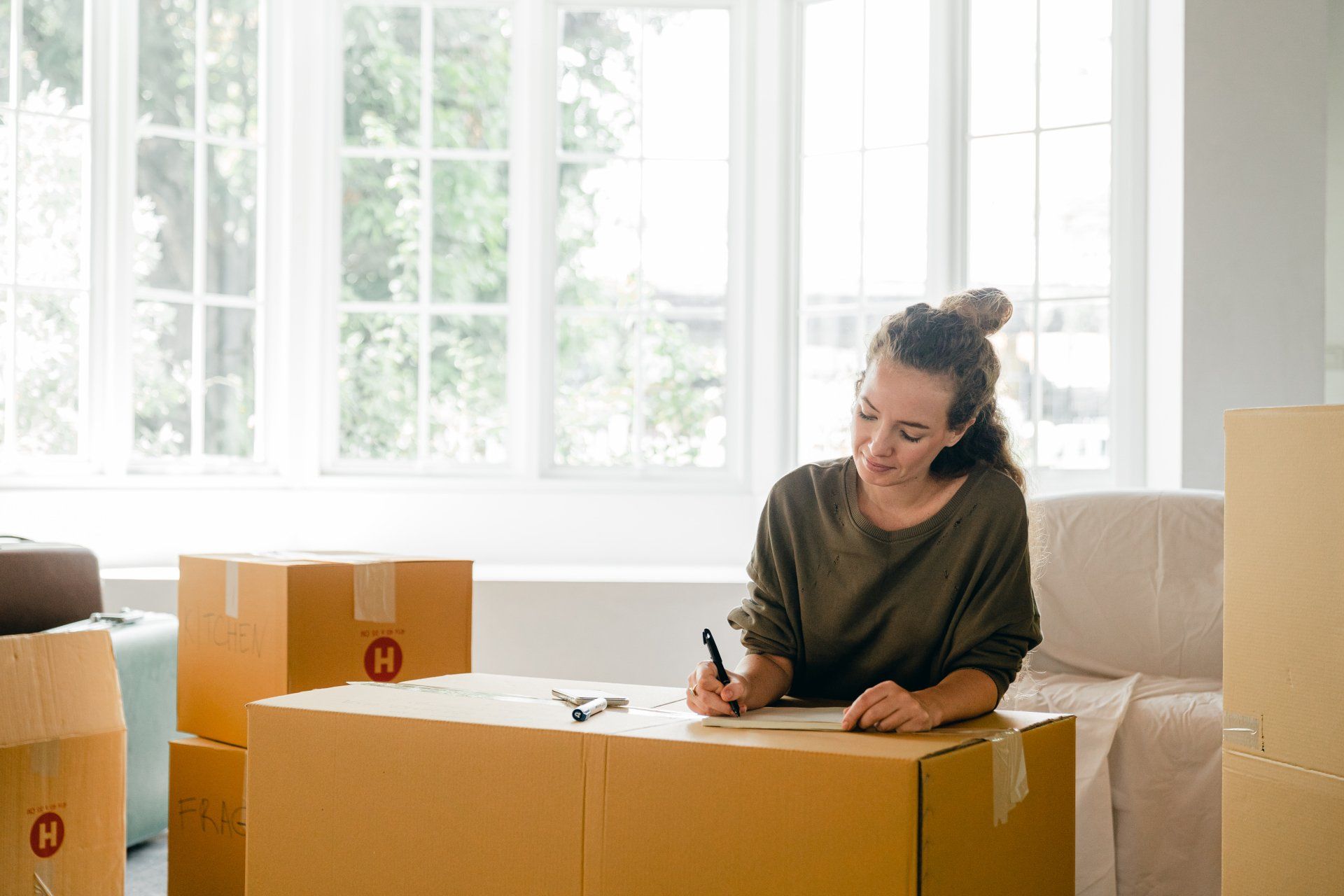 A woman is sitting on a cardboard box in a living room writing on a piece of paper.