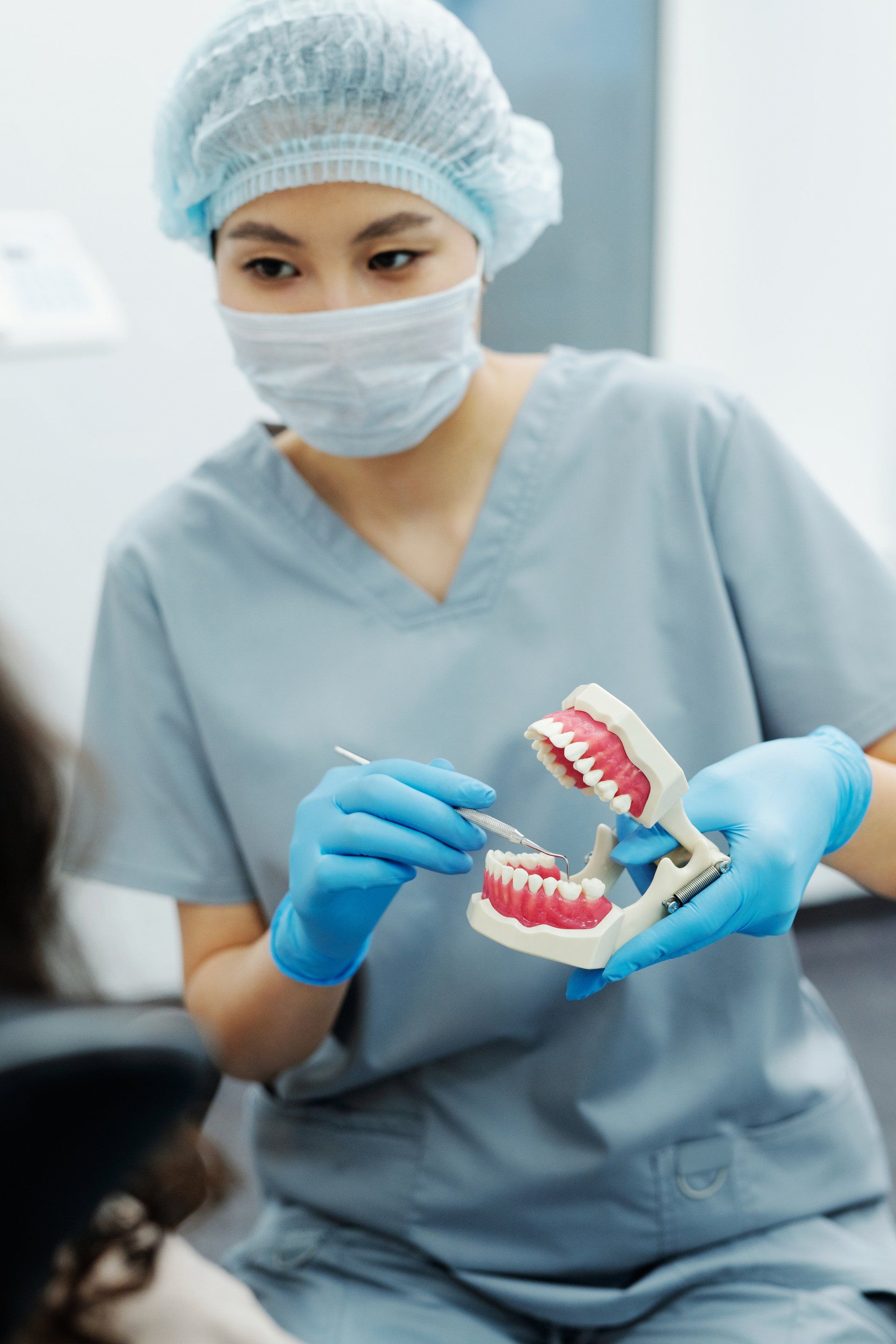 Dentist in scrubs, mask, and gloves, showing teeth model to patient.