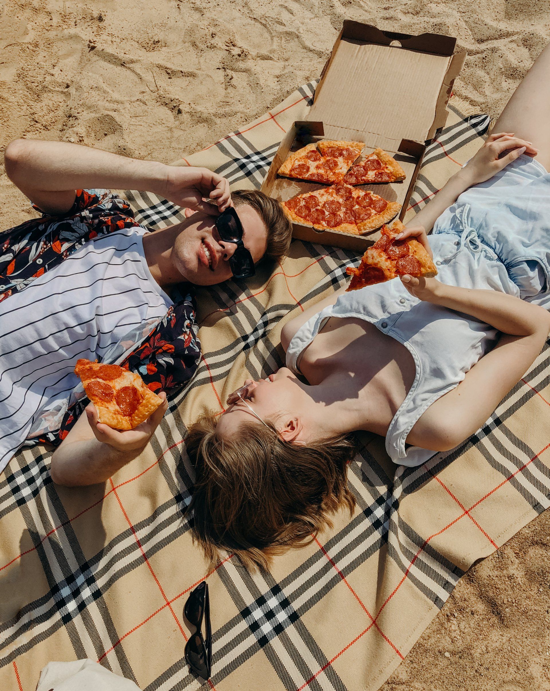 Un hombre y una mujer están tumbados sobre una manta en la playa comiendo pizza.