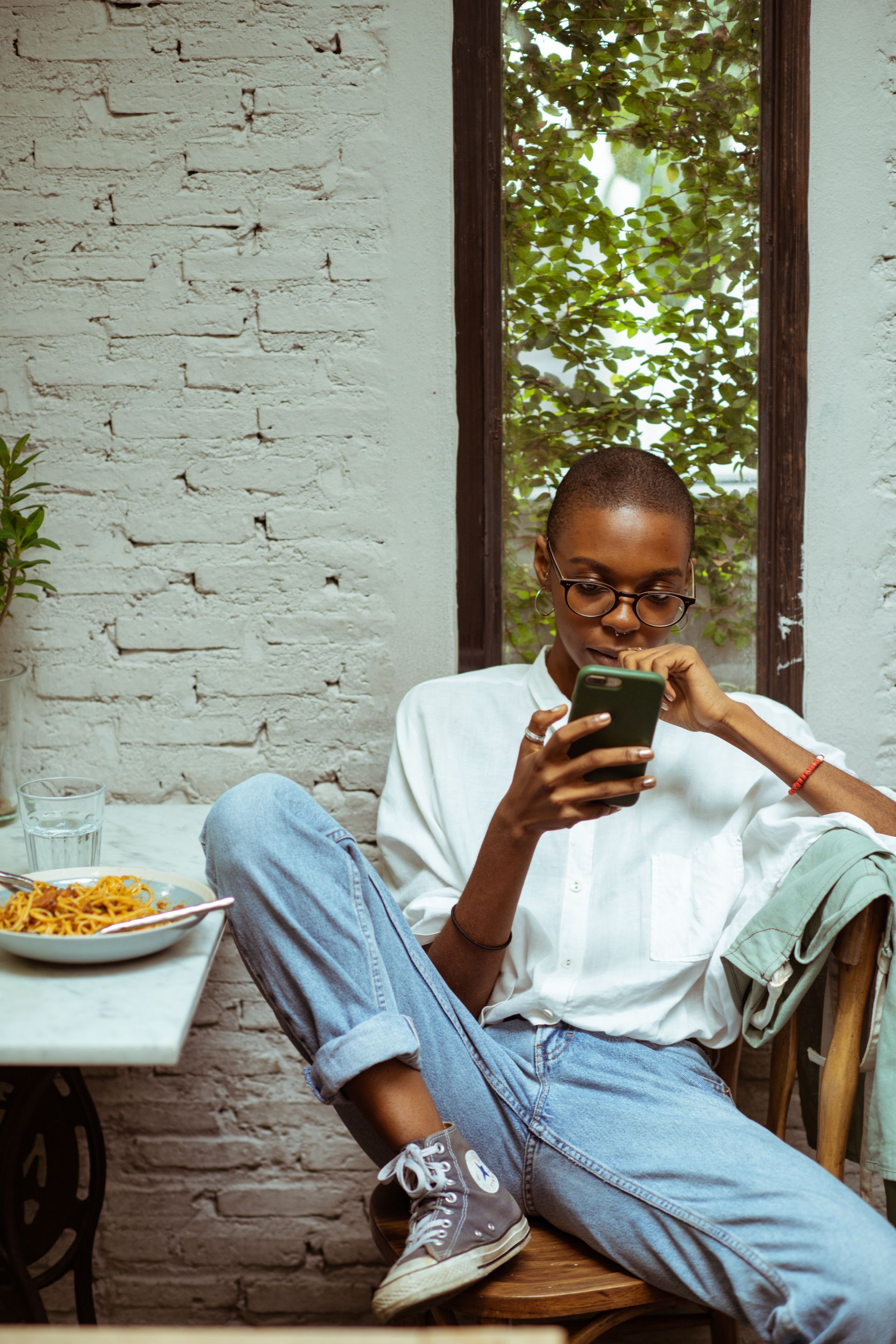 Woman in white shirt and jeans sits outdoors, looking at phone. Plate of food on table.