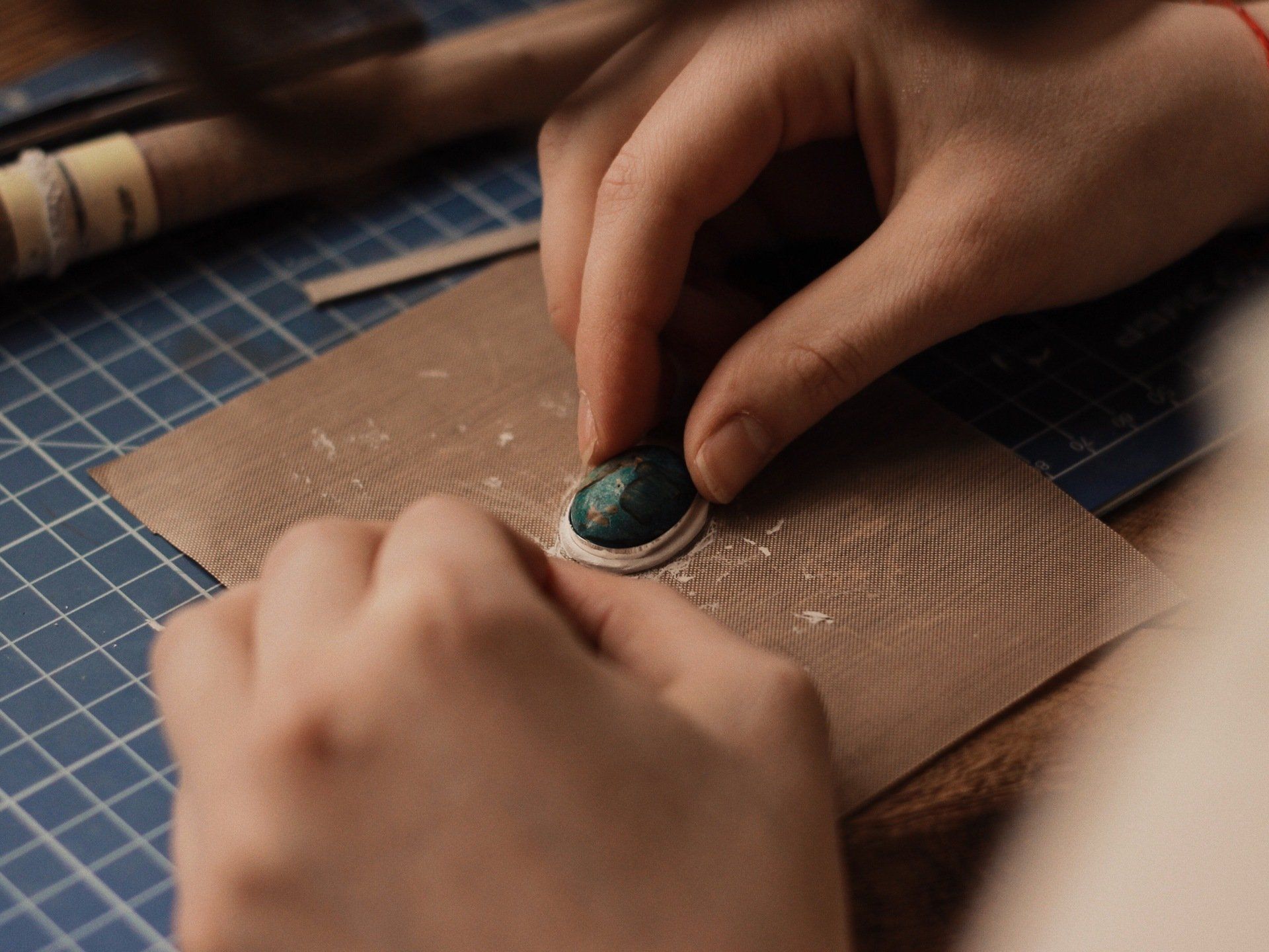 Hands crafting a pendant with a green gemstone and silver border on a blue grid mat.