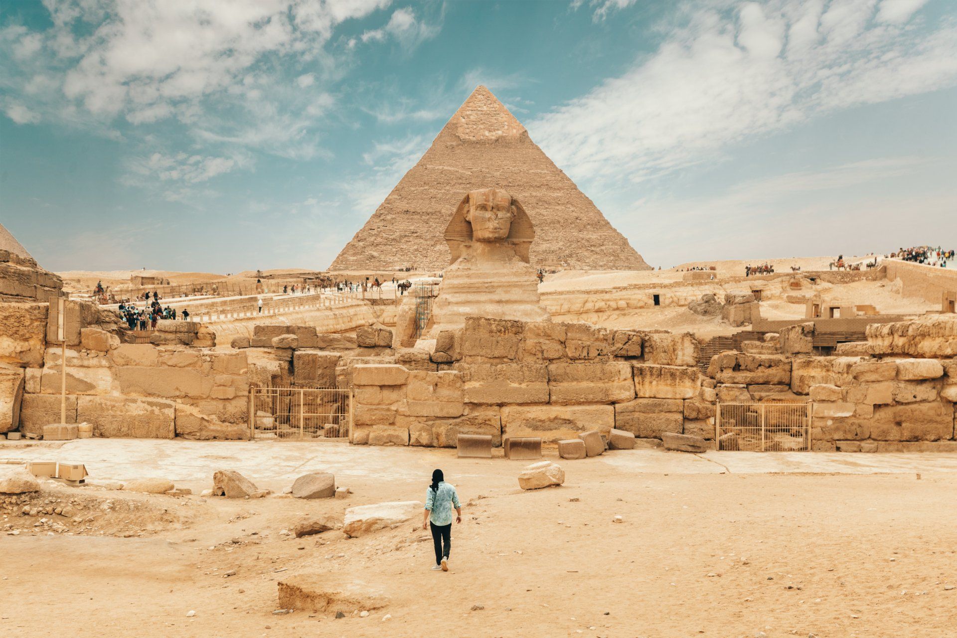 A woman is walking in front of a pyramid and sphinx in the desert.