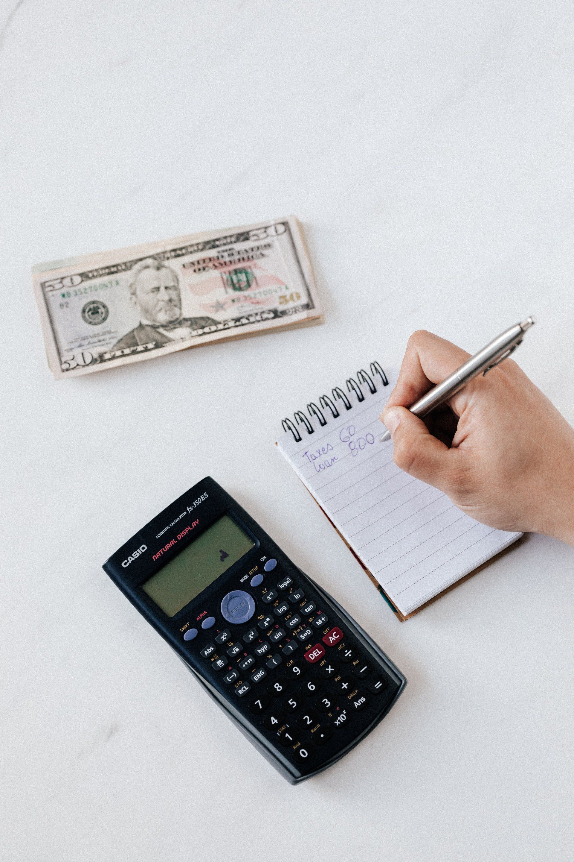 A person is writing in a notebook next to a calculator and a stack of money.