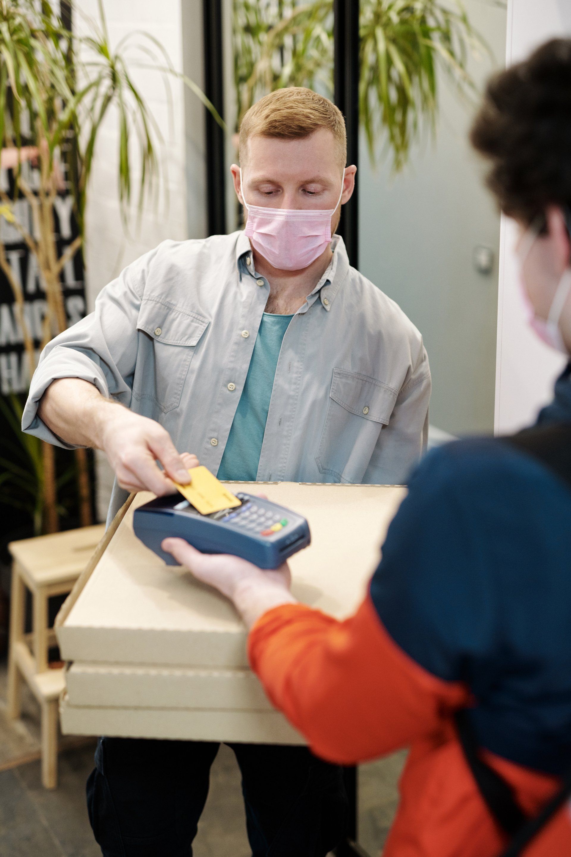 A man wearing a mask is holding a box of pizza and a credit card.
