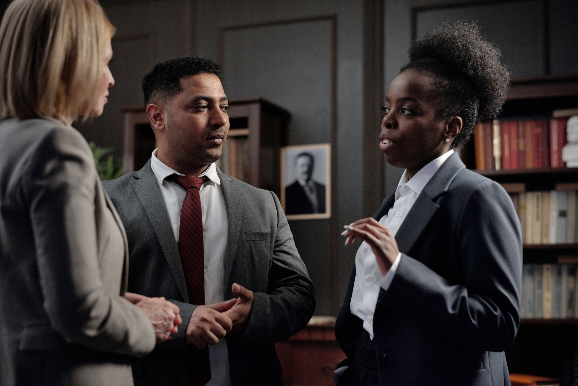 Three people in suits talking in a room with a bookshelf and a portrait.