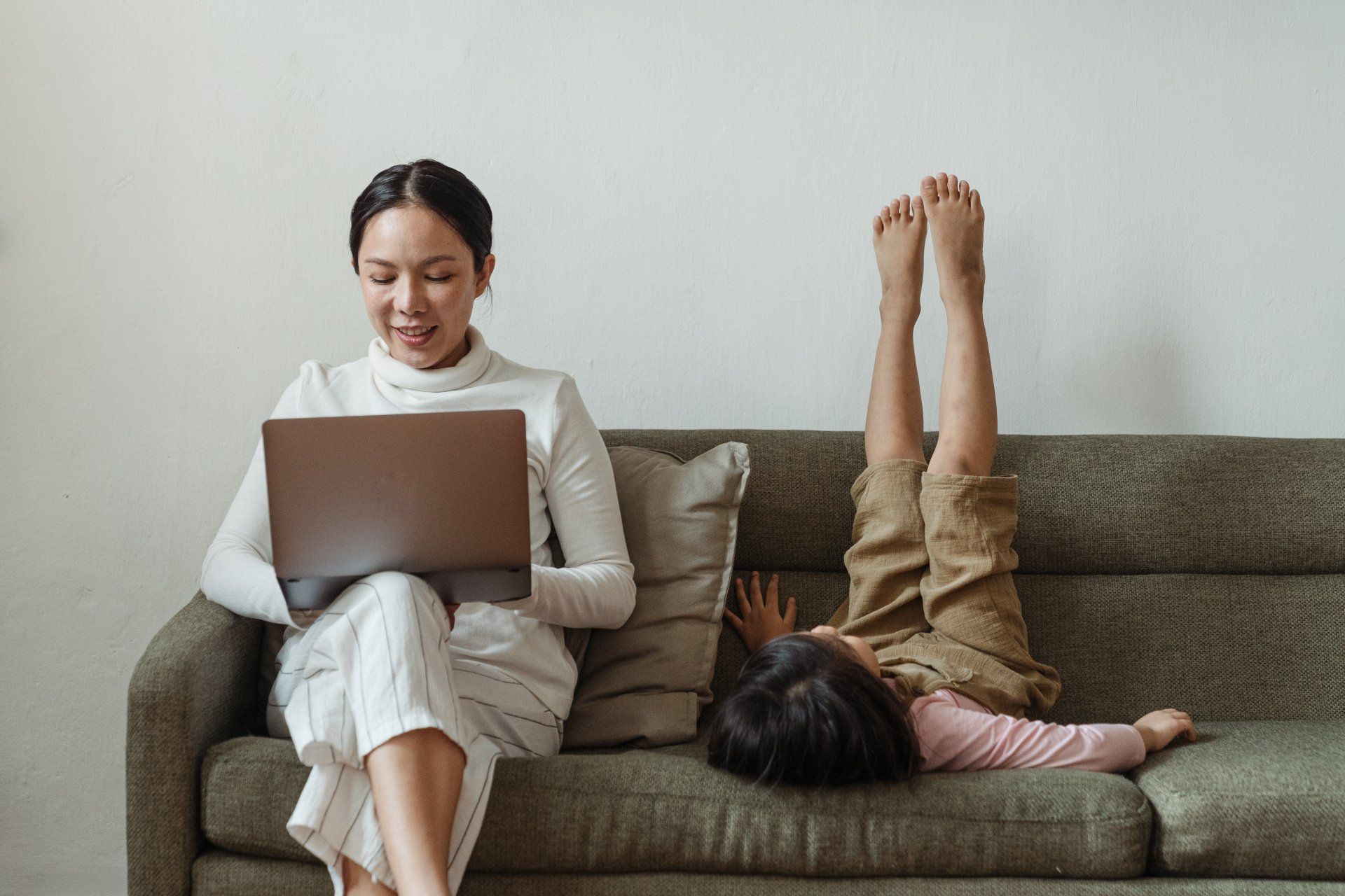 A woman is sitting on a couch using a laptop and a child is laying on the couch.