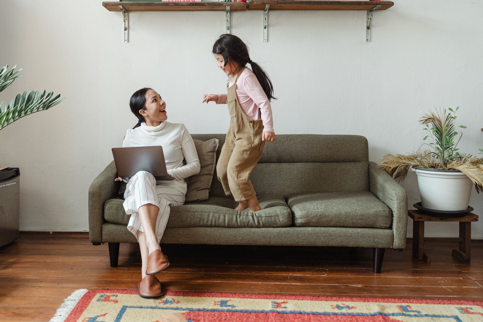 A woman is sitting on a couch with a laptop and a little girl is jumping on the couch.