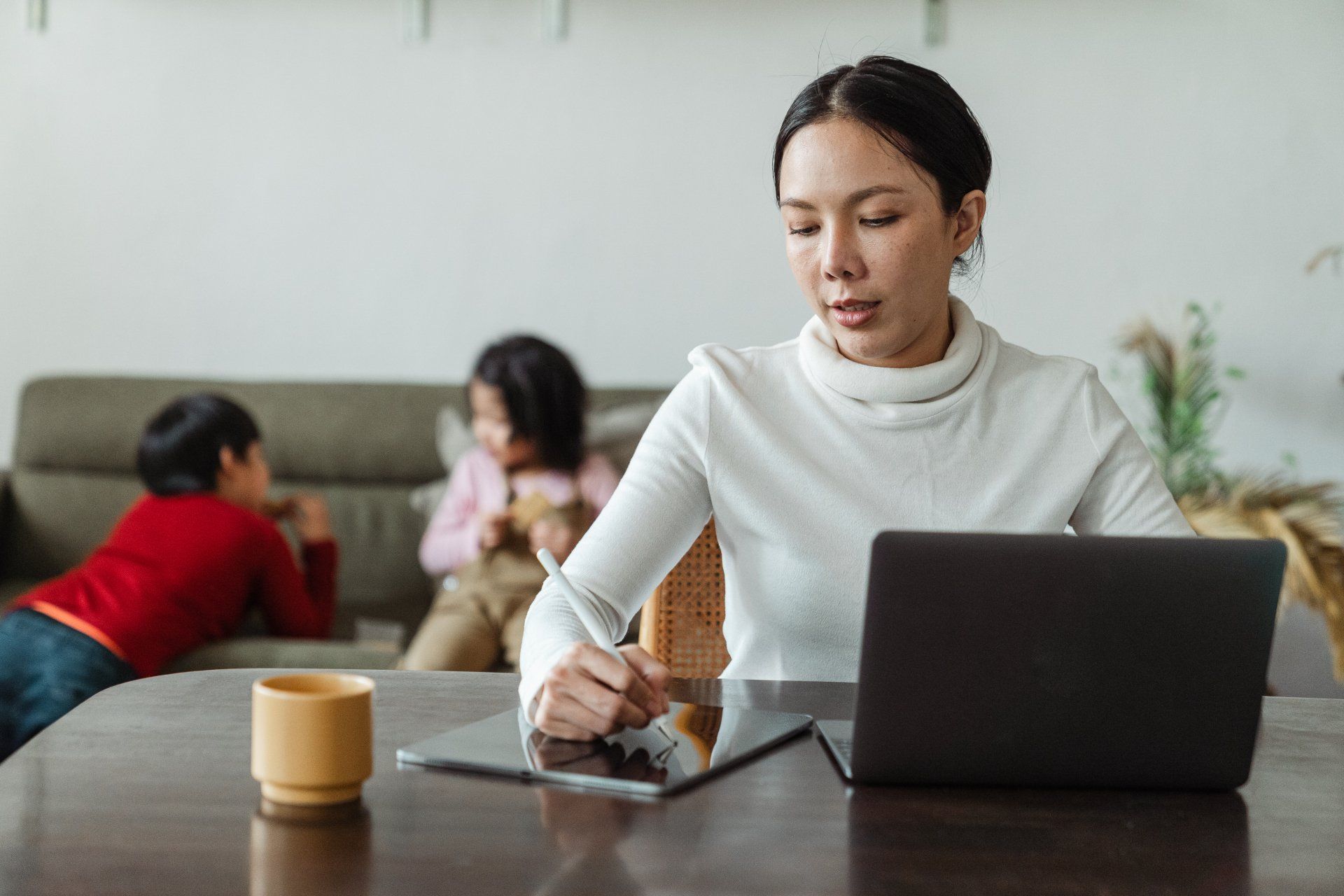 Donna che lavora al computer portatile e al tablet al tavolo, sullo sfondo bambini che giocano sul divano.