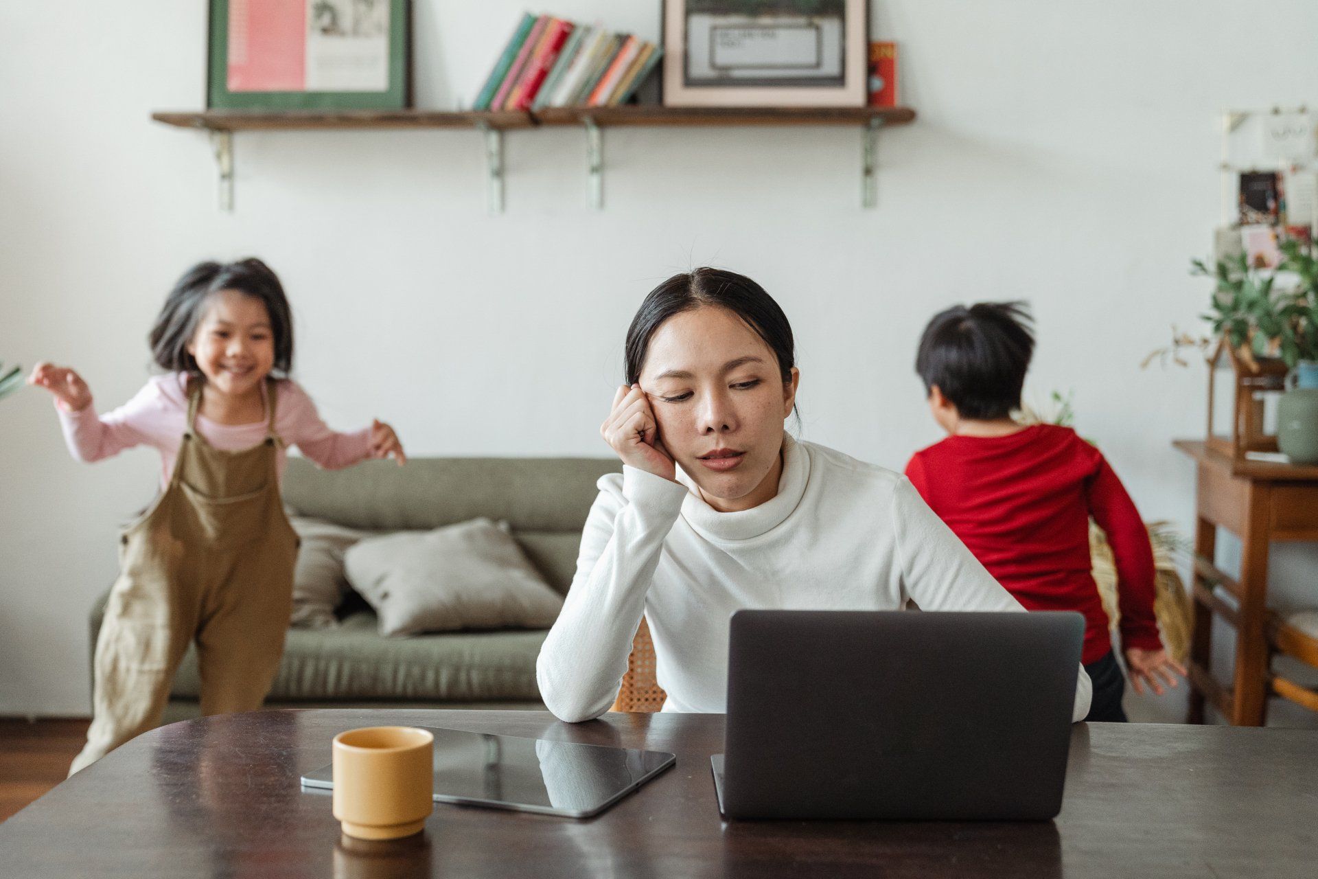 a woman sits at a table with a laptop and two children playing in the background