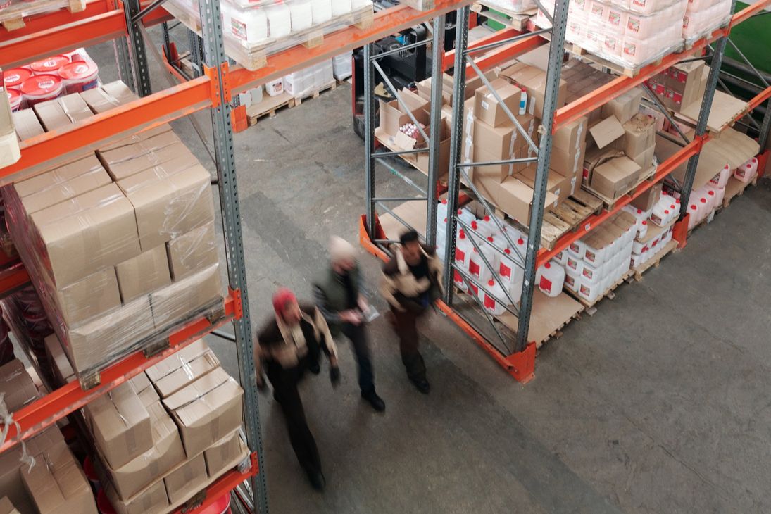 Tre operai in un corridoio di un magazzino, tra scatole impilate e scaffalature per pallet, guardano verso l'alto.