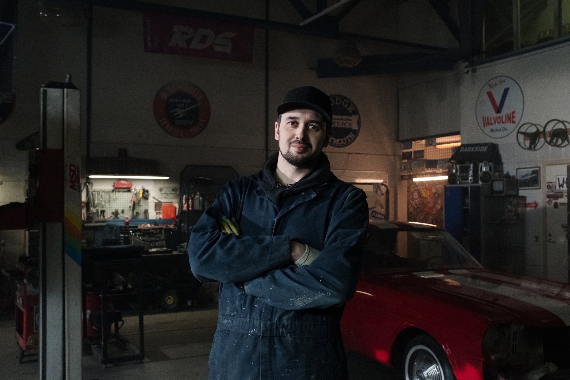 Mechanic in blue overalls stands with arms crossed in a garage next to a red sports car.