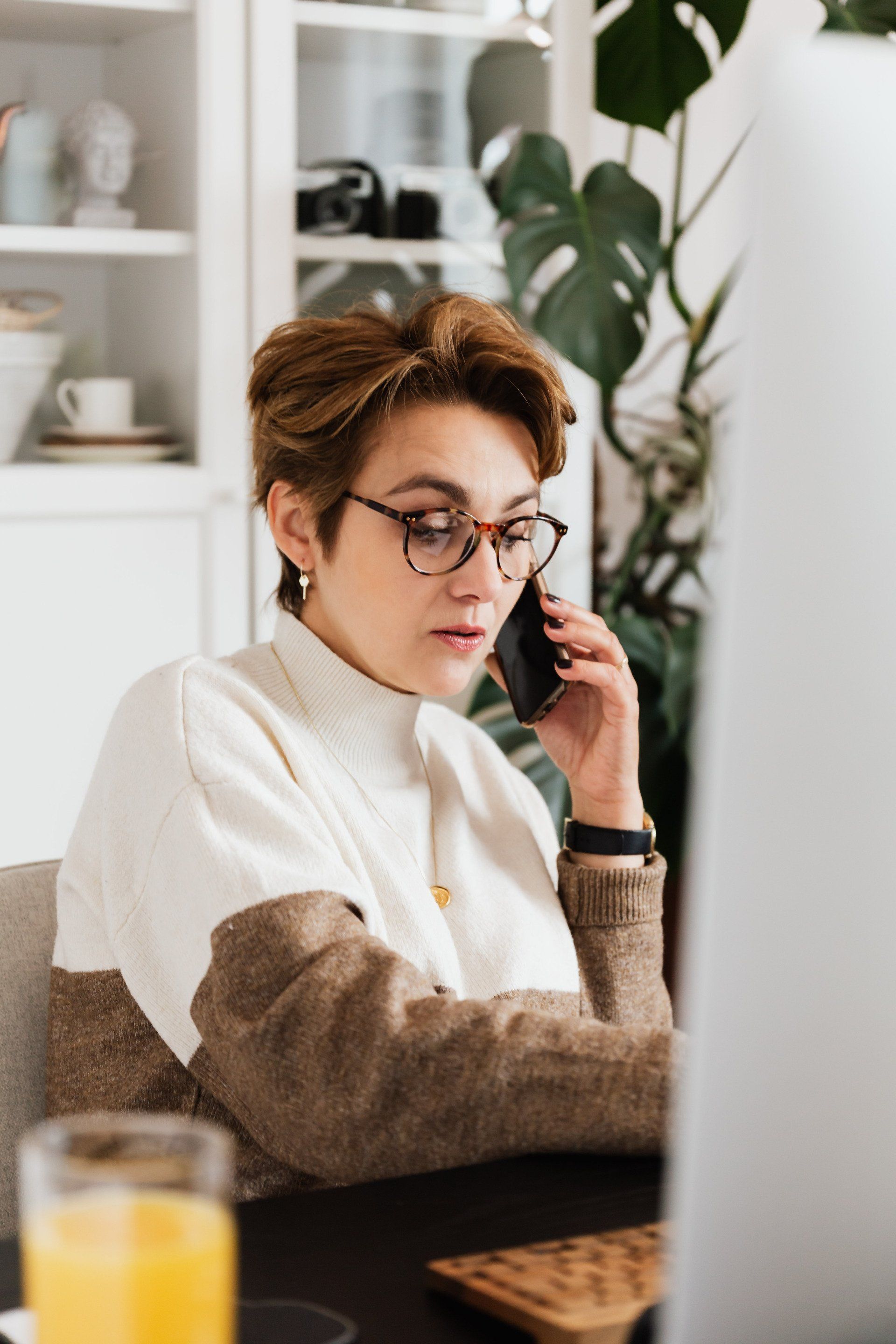 A woman is sitting at a table talking on a cell phone in front of a laptop computer.