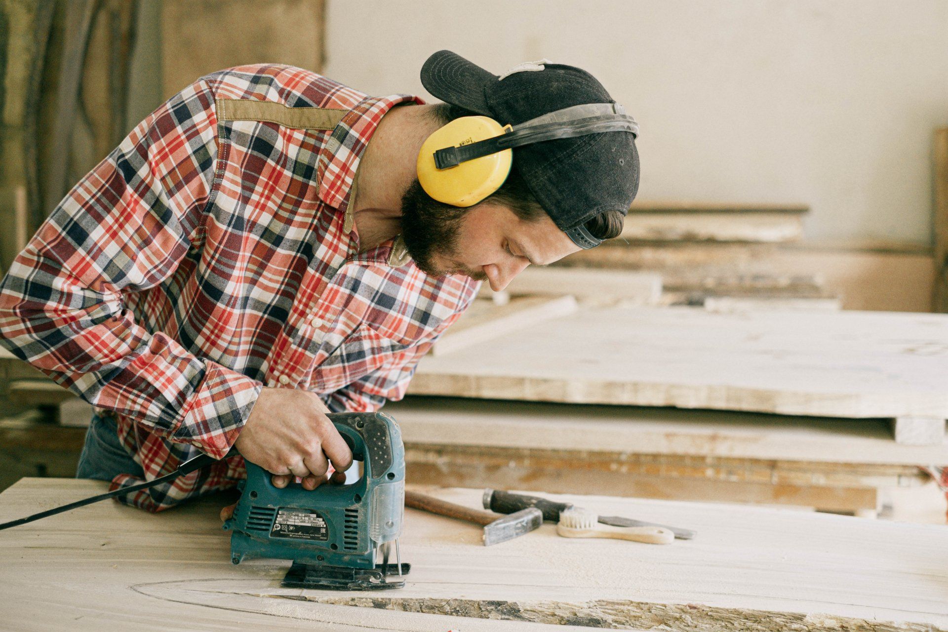Ocean County Handyman using a jigsaw on wood, wearing ear protection and a plaid shirt, in a workshop.