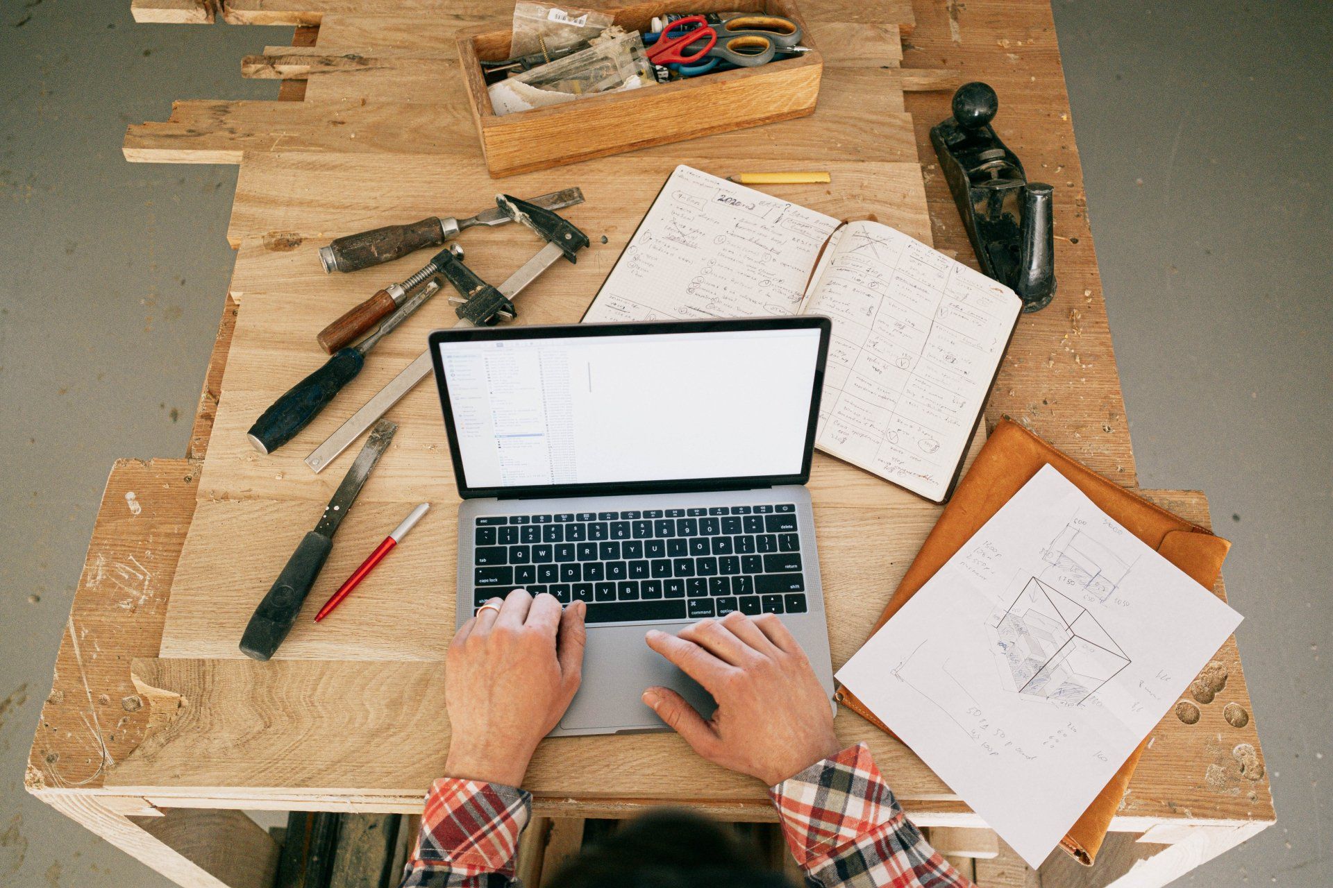 Hands typing on laptop at a cluttered wooden desk with tools, notebooks, and sketches