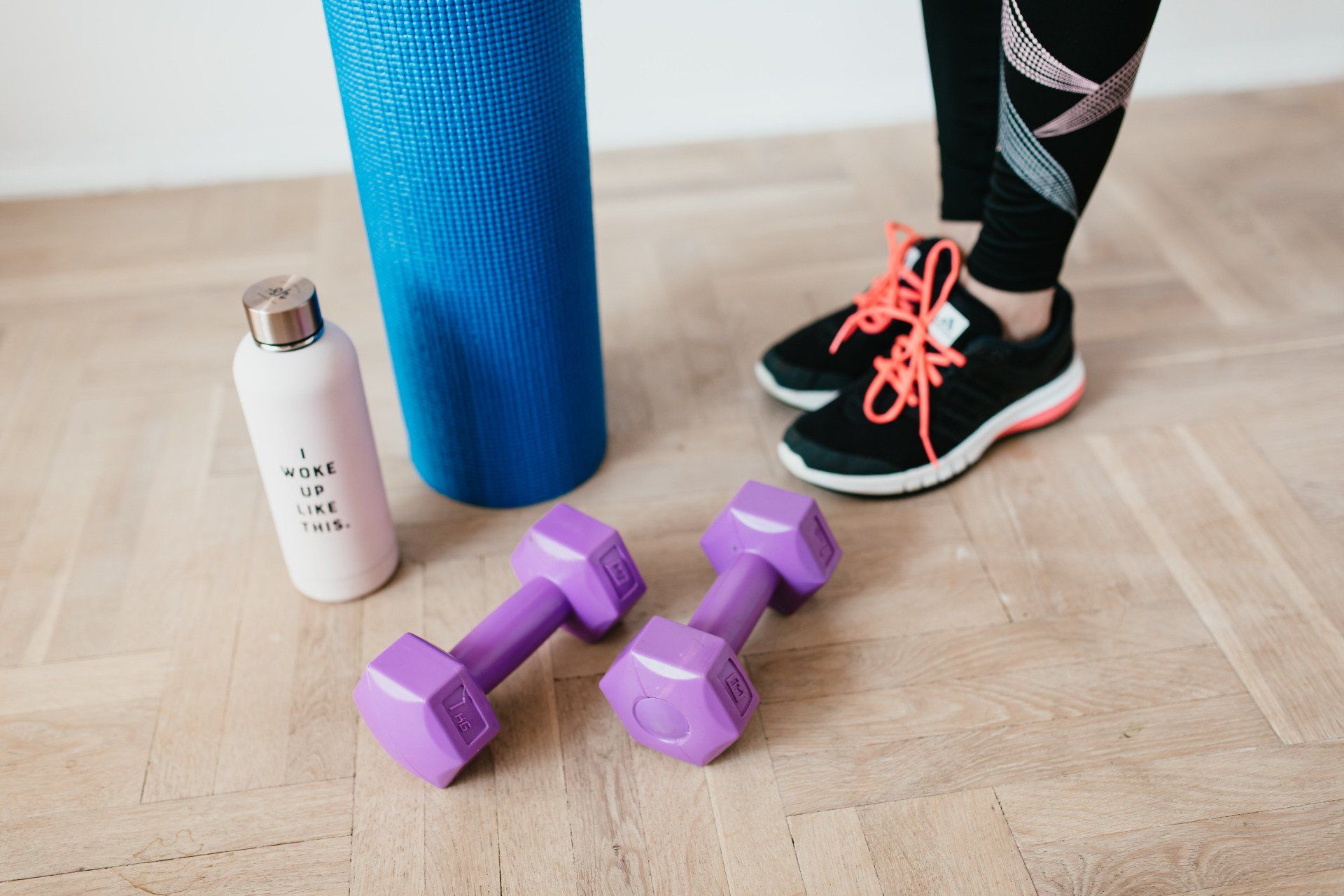 A woman is standing next to a pair of purple dumbbells and a yoga mat.
