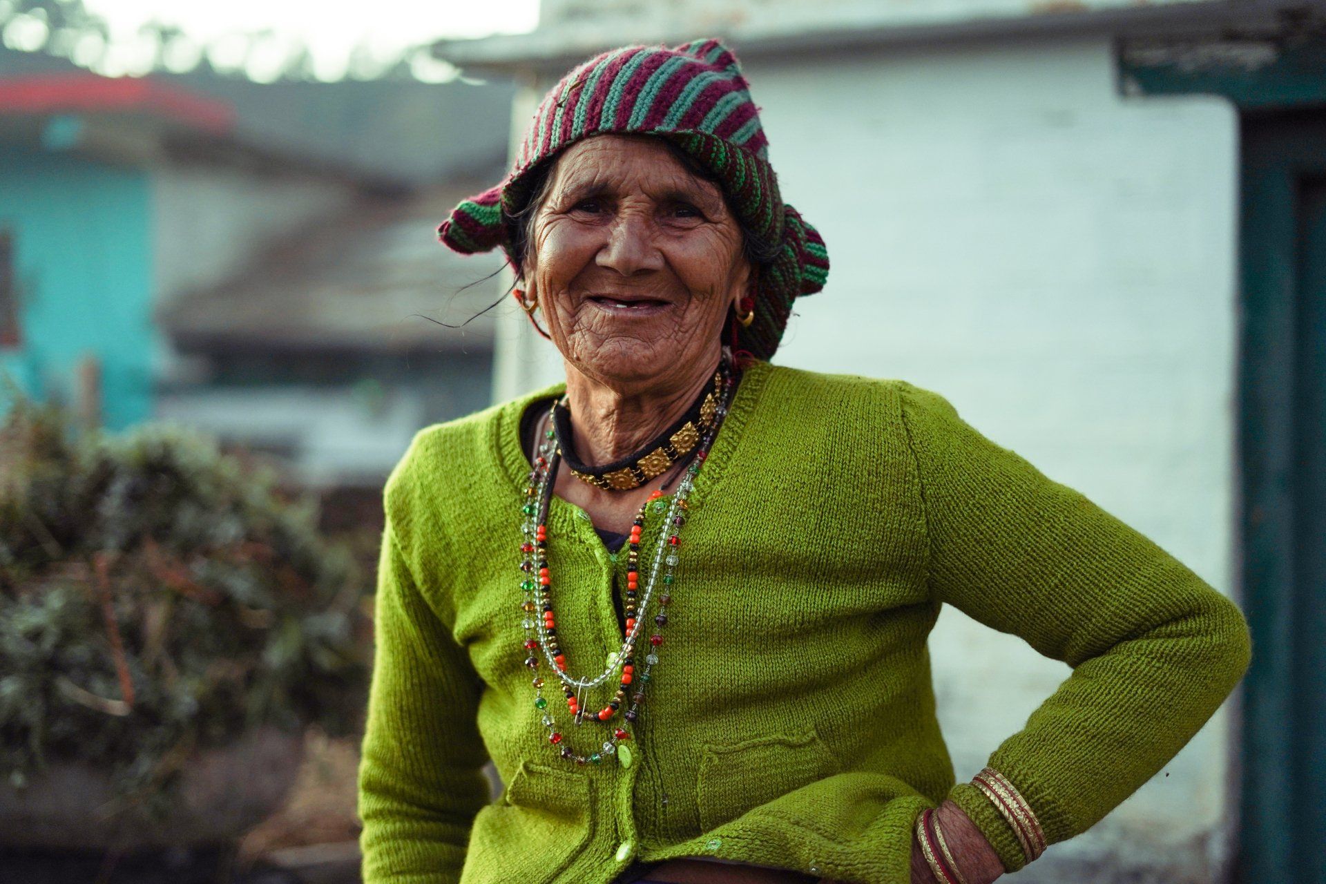 An elderly woman wearing a green sweater and a hat is standing with her hands on her hips.