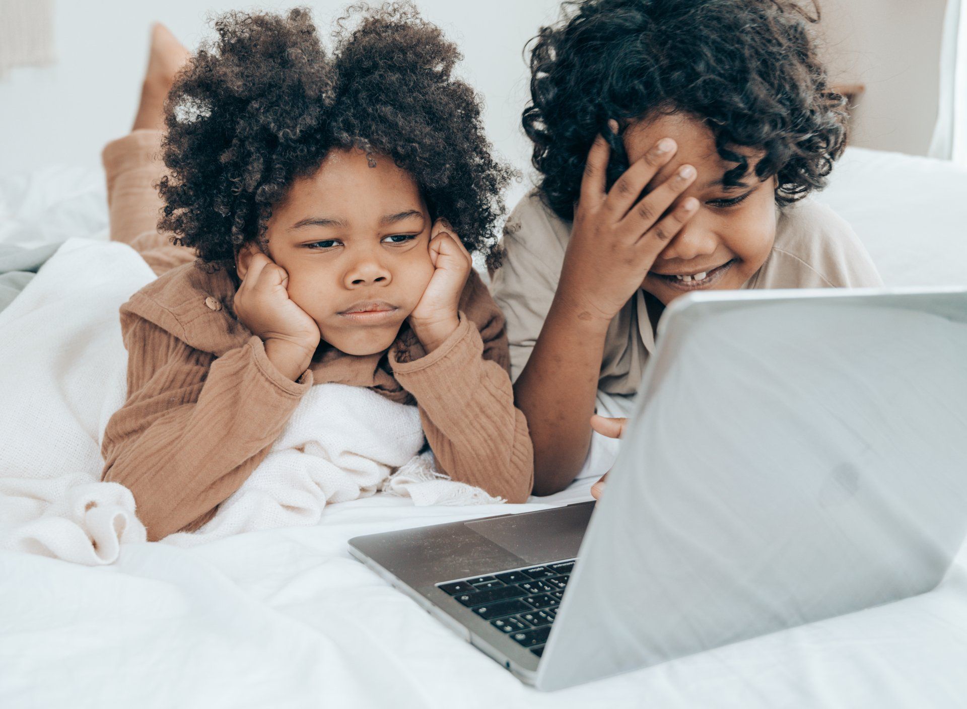 Two children are laying on a bed looking at a laptop computer.