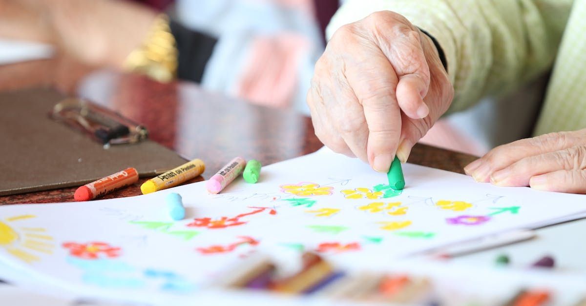 Person's hands using a green crayon to color on a white paper with other colorful crayons, close-up.