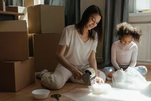 A person taping a cardboard box while a child sits nearby on the floor wrapping items in bubble wrap for moving.