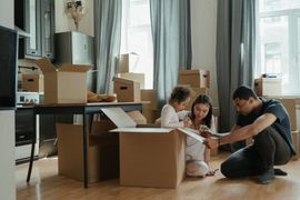 Family unpacking boxes in a new home: child, parents, cardboard boxes, light interior, excited expressions.