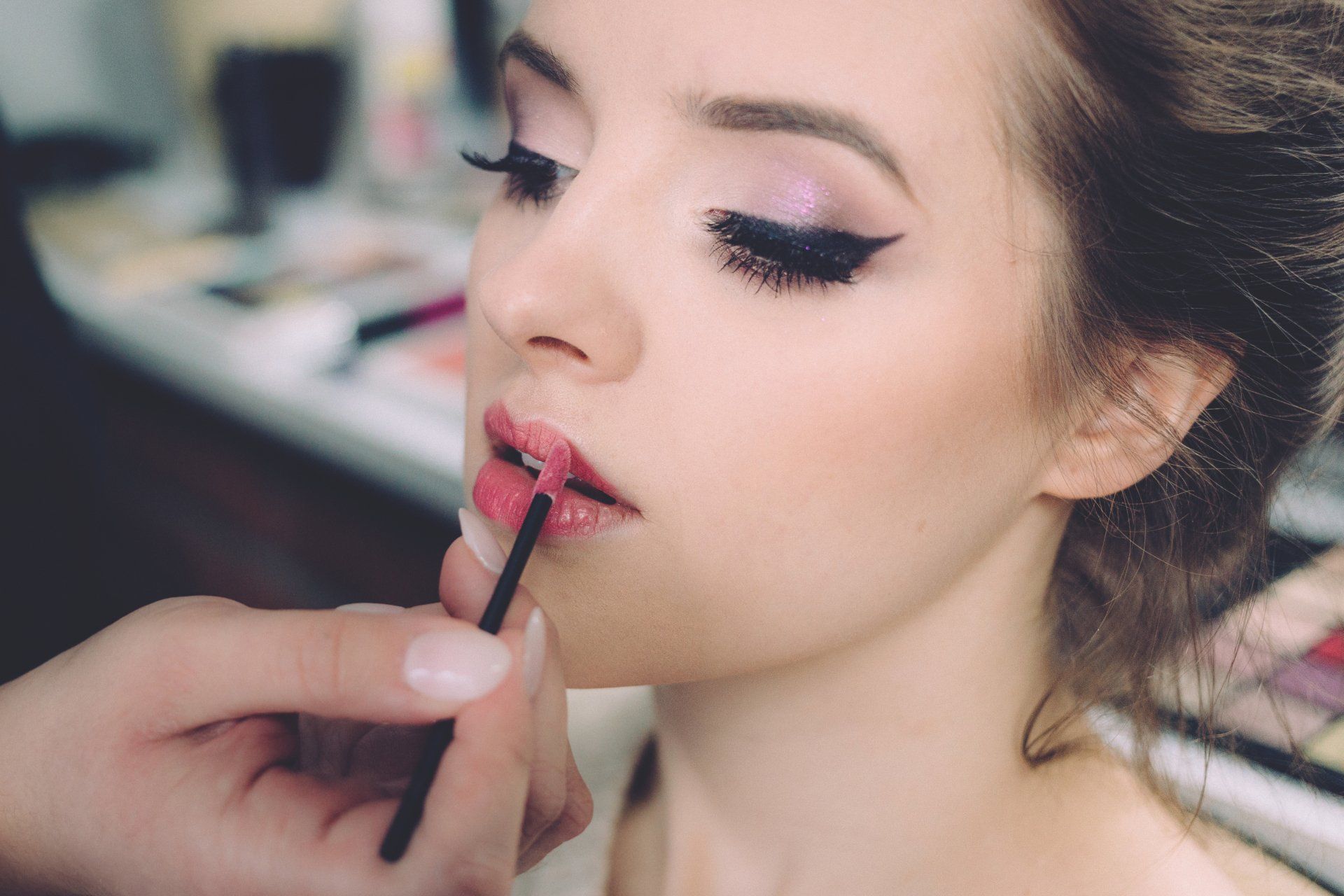 A makeup artist uses a brush to apply pink lip gloss to a person’s lips in a studio setting.