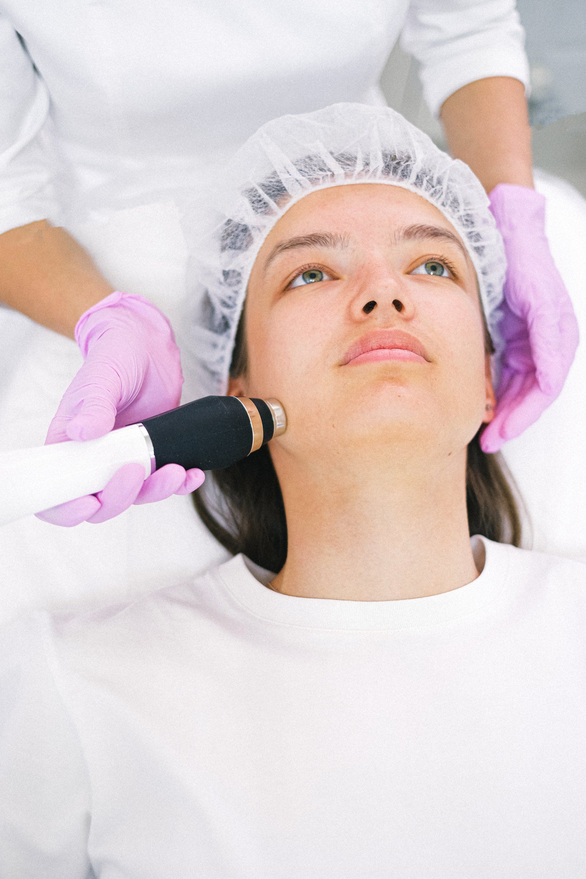 Women Laying down in a surgical cap another woman wearing gloves touching laser pen near her jawline