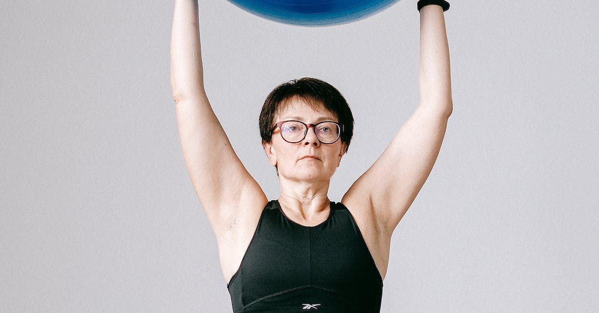 Mujer sosteniendo una pelota de ejercicio azul sobre su cabeza, con gafas y una camiseta sin mangas negra, en un estudio.