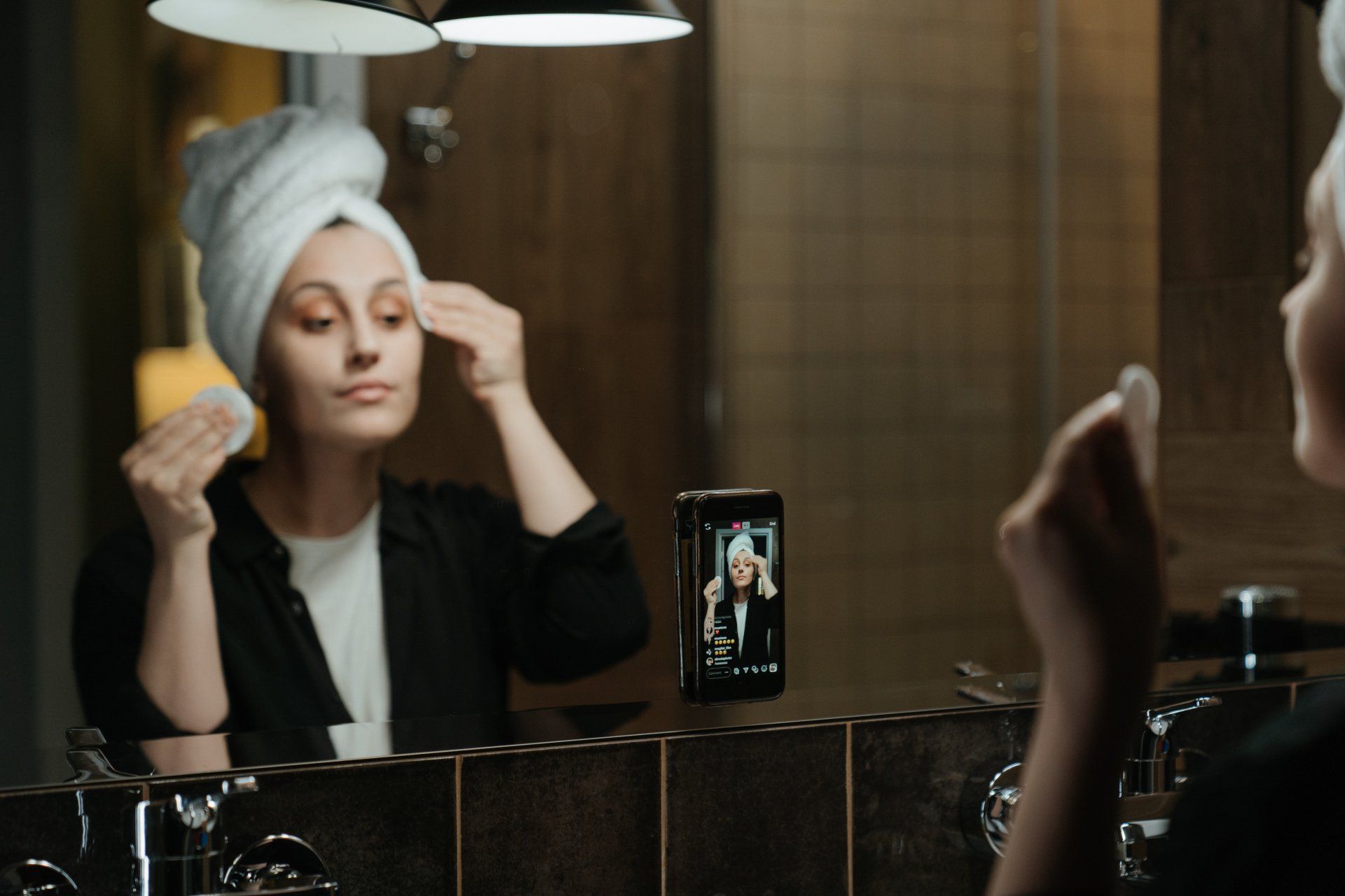 A woman with a towel wrapped around her head is looking at her face in a bathroom mirror.