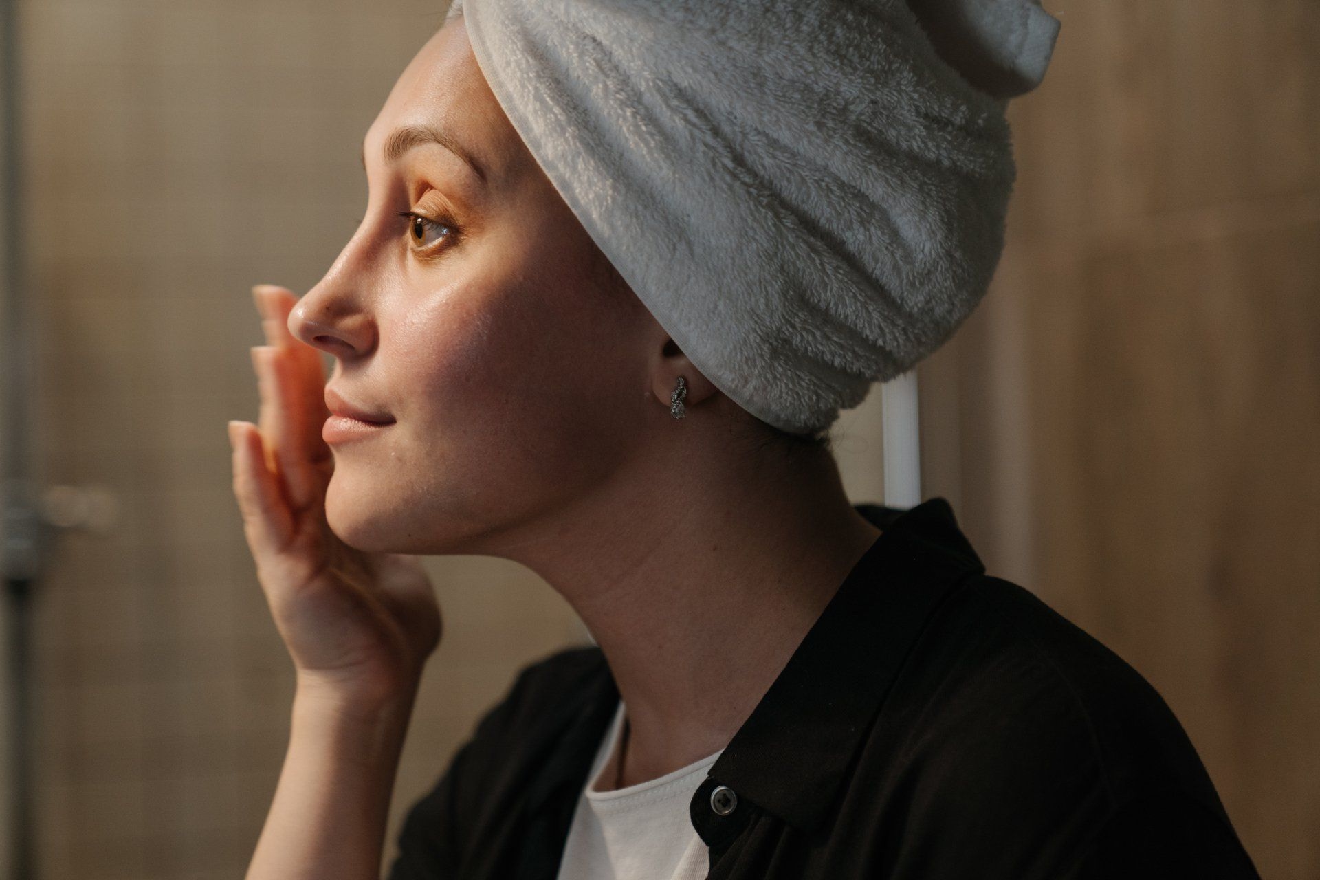 Woman with towel on head, applying product to face, in bathroom.