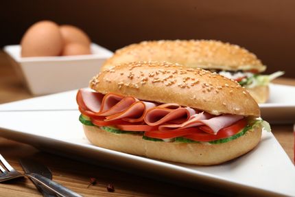 Een subsandwich met ham, tomaten, sla en komkommer op een witte plaat.