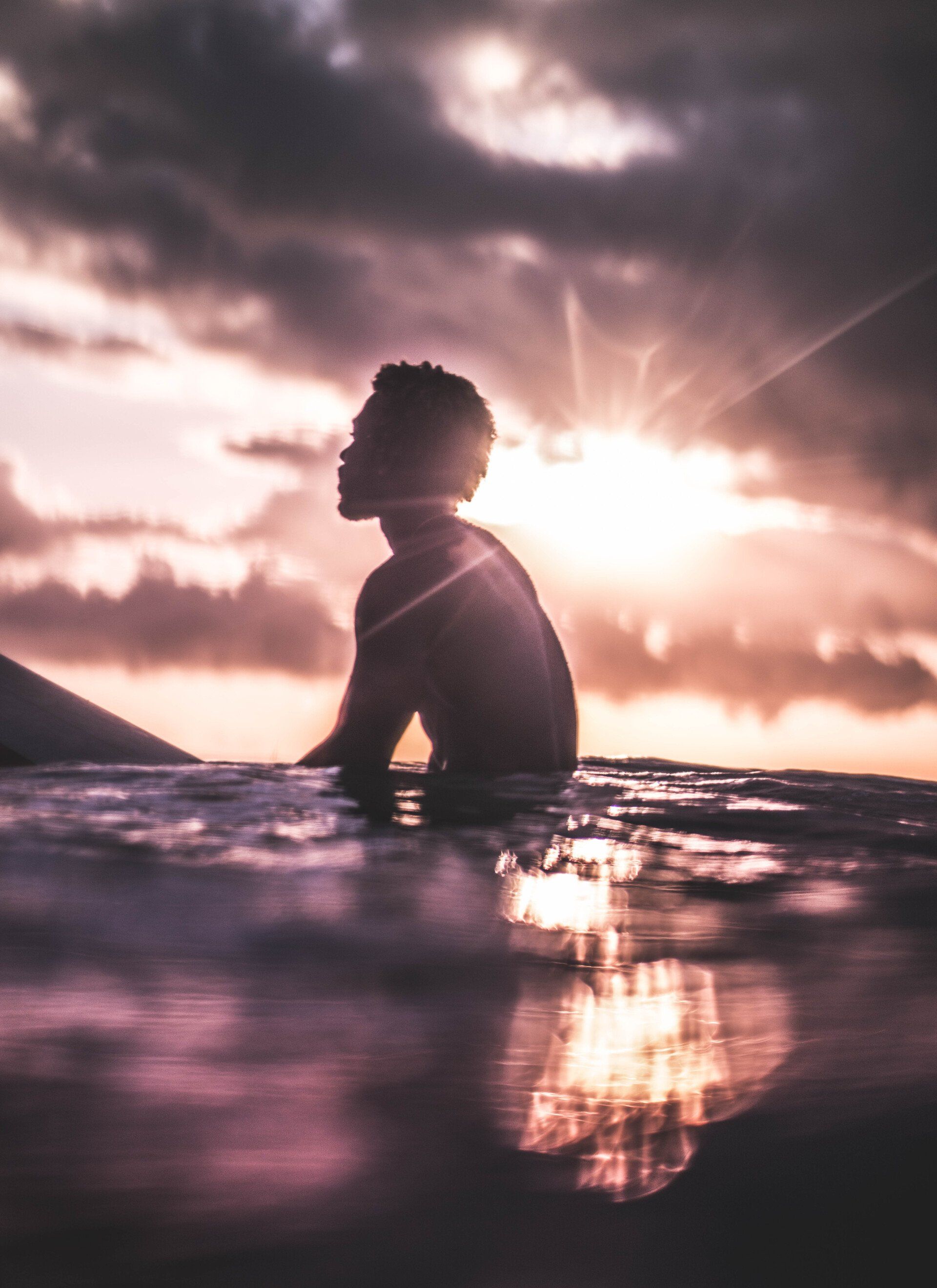 Person on surfboard in water, silhouetted against a cloudy sunset; sun reflecting in the water.