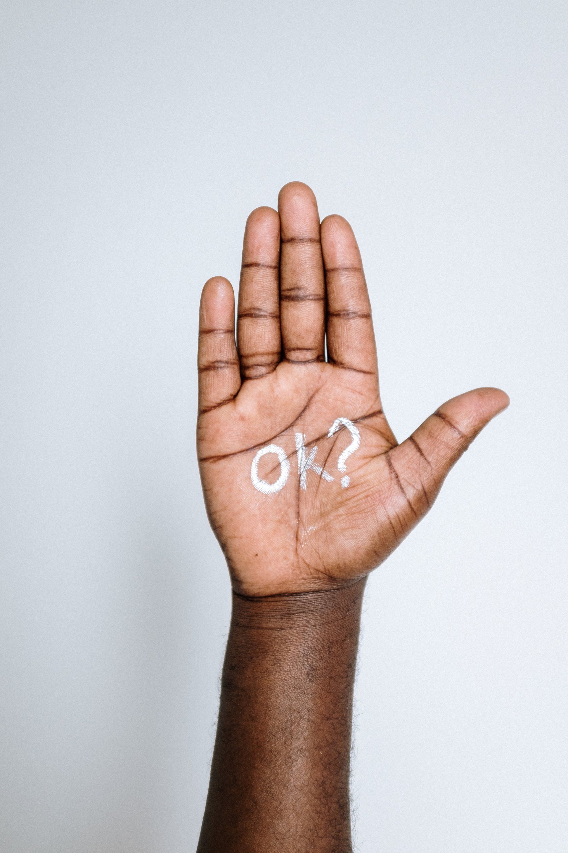 Paume ouverte avec le mot « OK ? » écrit en blanc. Peau brune sur fond blanc.