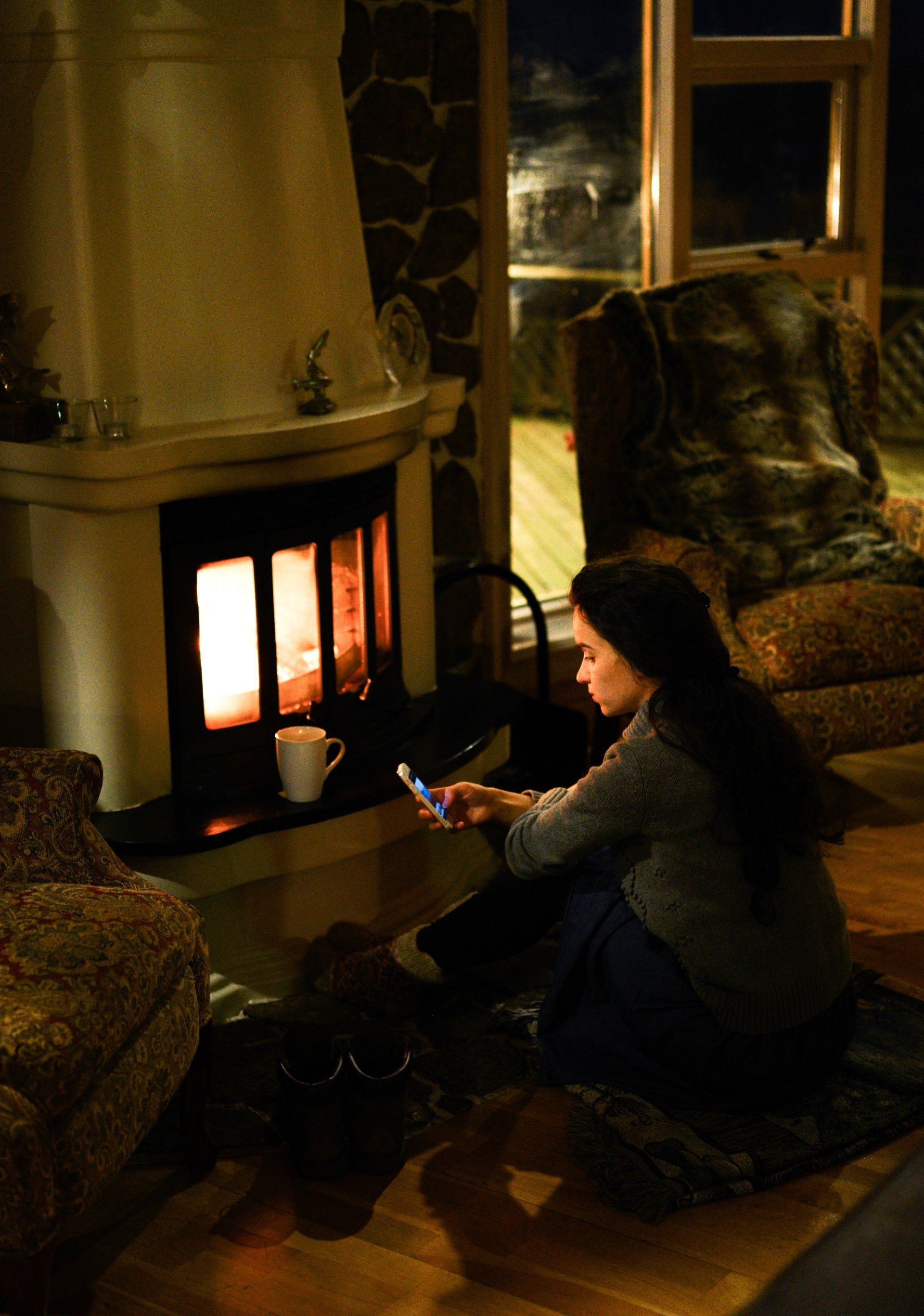 A woman is sitting in front of a fireplace looking at her phone.