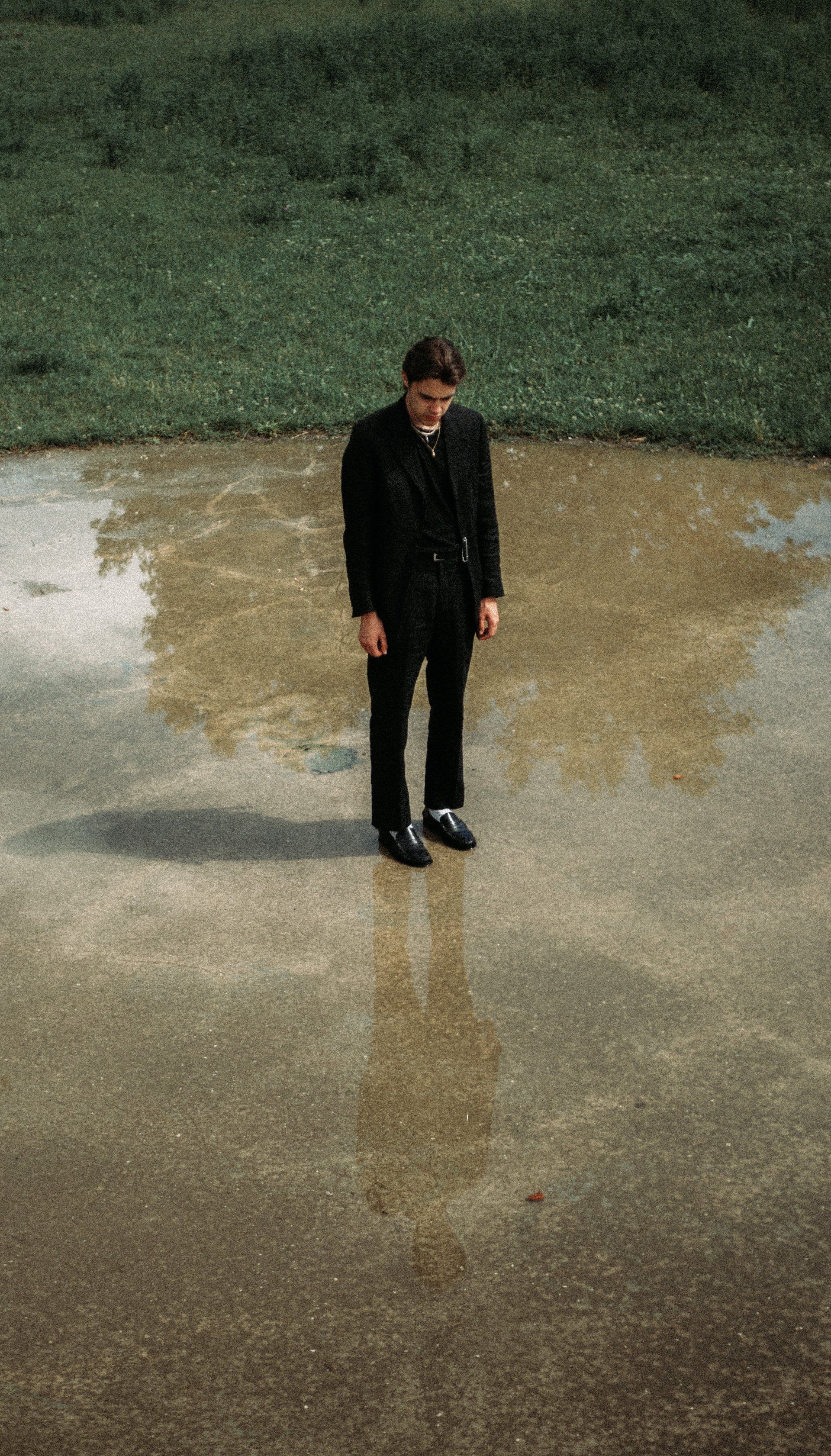 Hombre con traje negro de pie en un charco, con reflejo visible. Hierba verde al fondo.