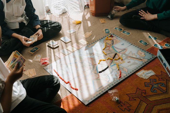 People gathered around a board game on the floor, with cards, tokens, and dice spread across a white mat.
