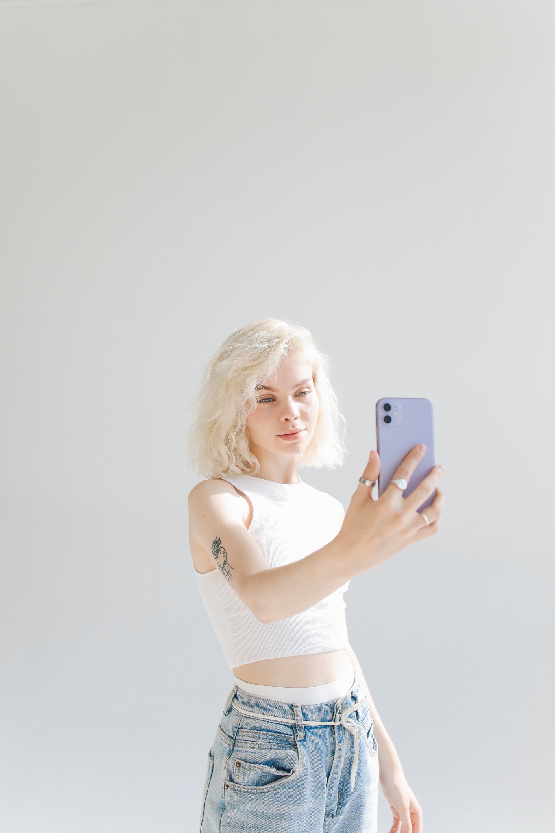 Image of a white woman with blonde hair and tattoo on her arm, holding a phone as if taking a selfie.