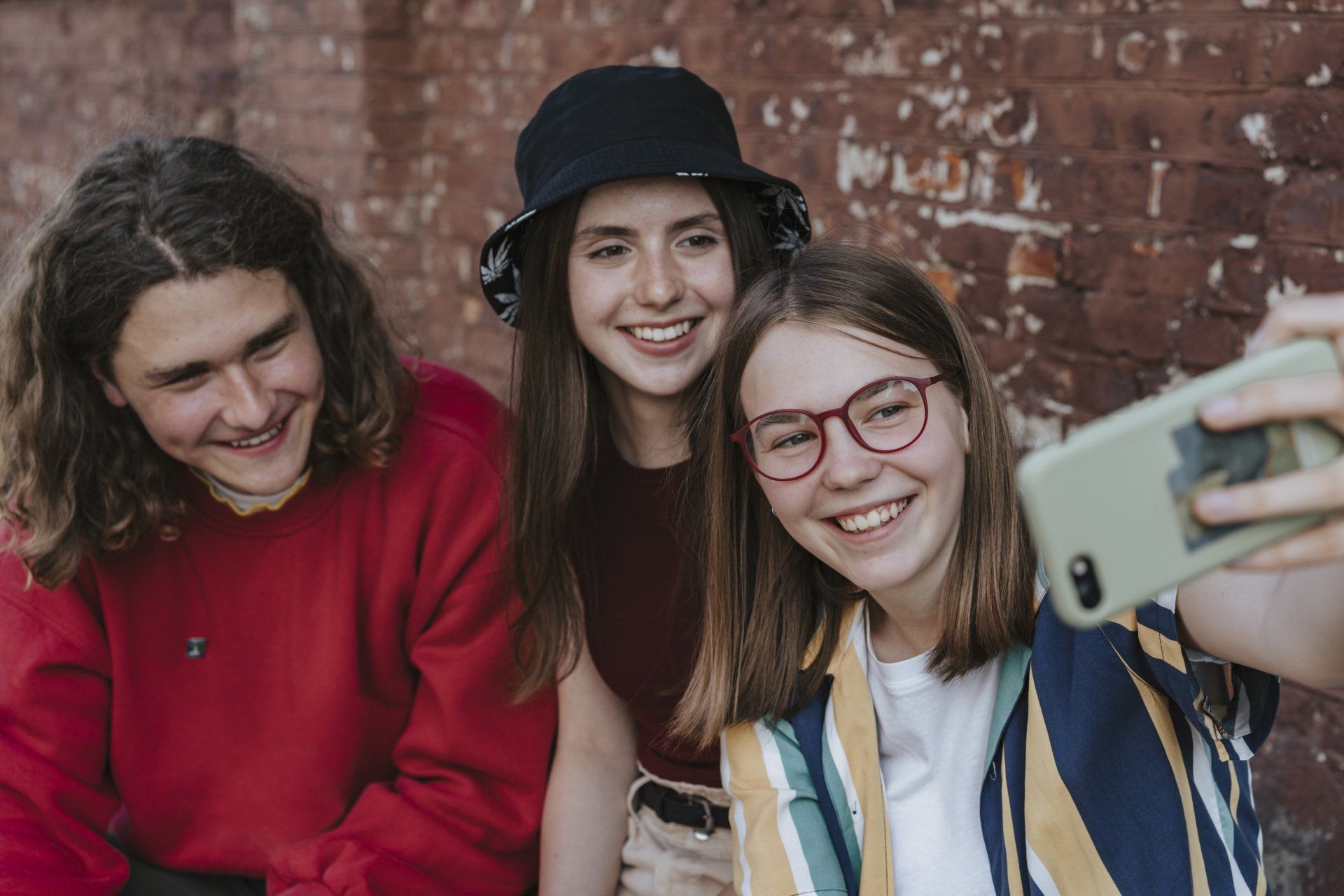 Three smiling people taking a selfie against a brick wall. One holds the phone.