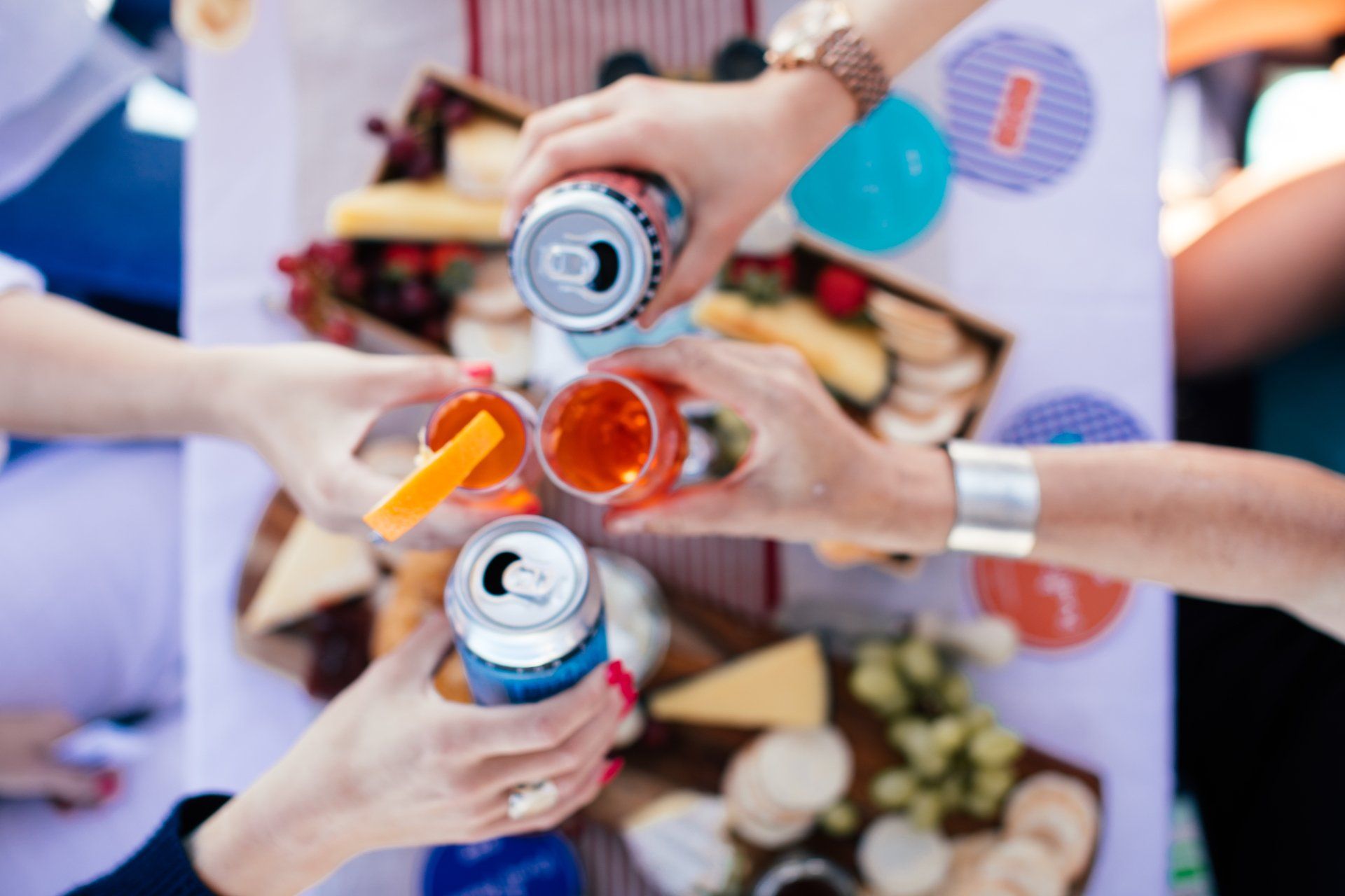 Hands toasting drinks over a picnic spread with cheese, crackers, and fruit.