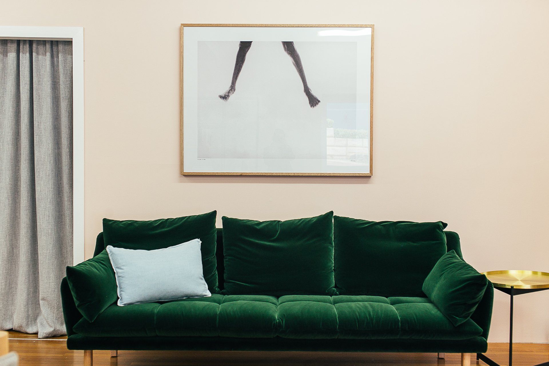 Emerald green sofa with a light blue pillow, below a framed art print. Light pink wall.