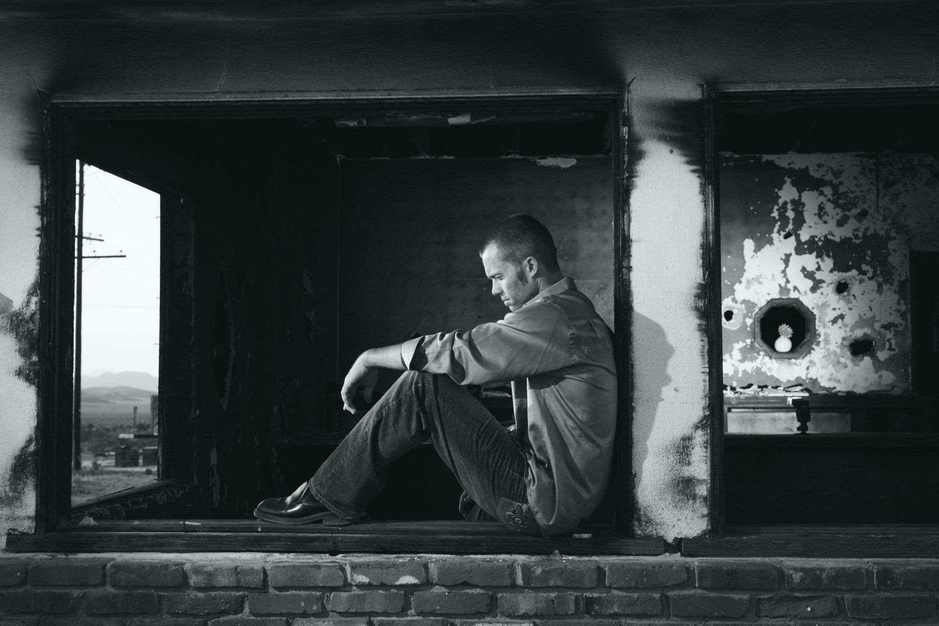 Man sits alone, head down, in a weathered doorway. Black and white.
