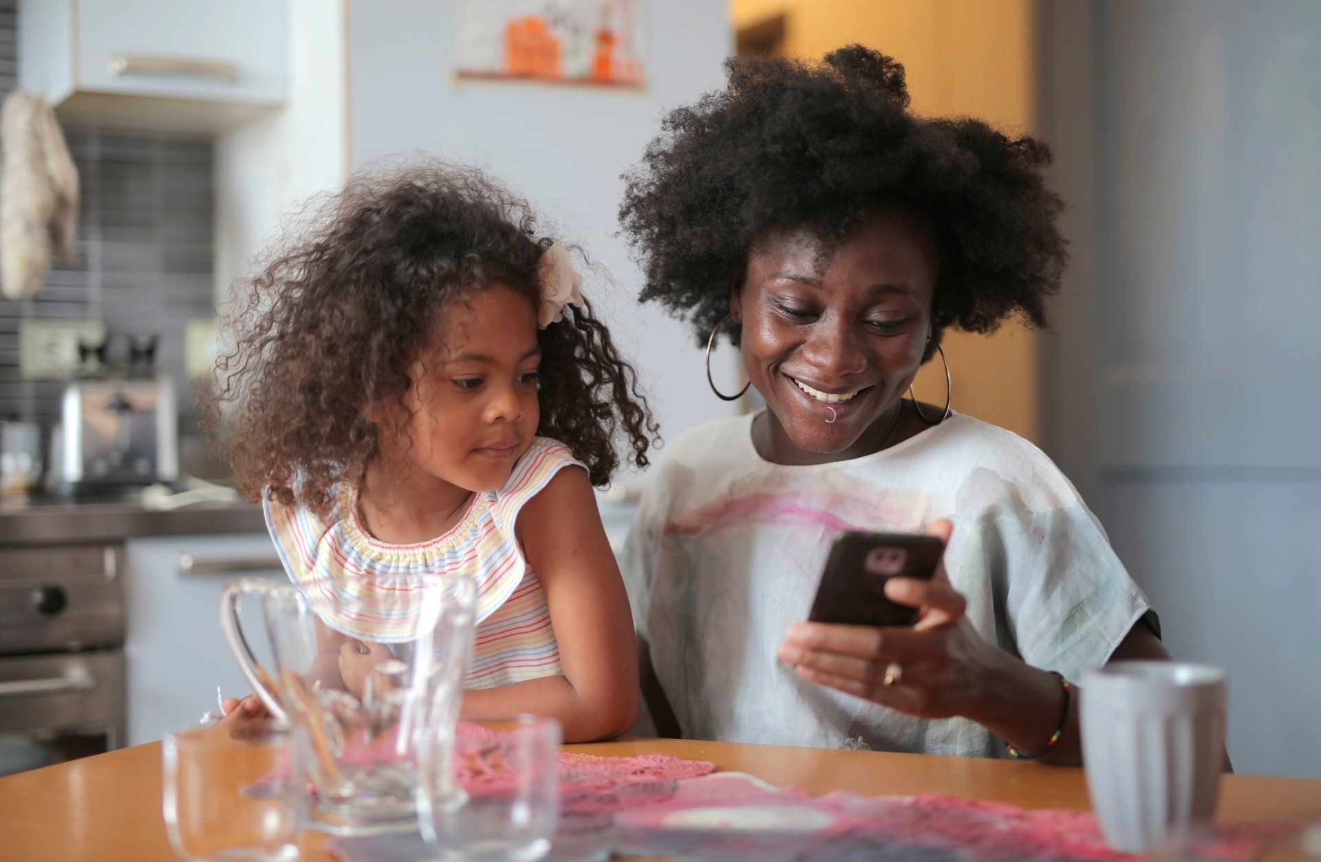 Woman and child at a kitchen table looking at a phone, smiling.