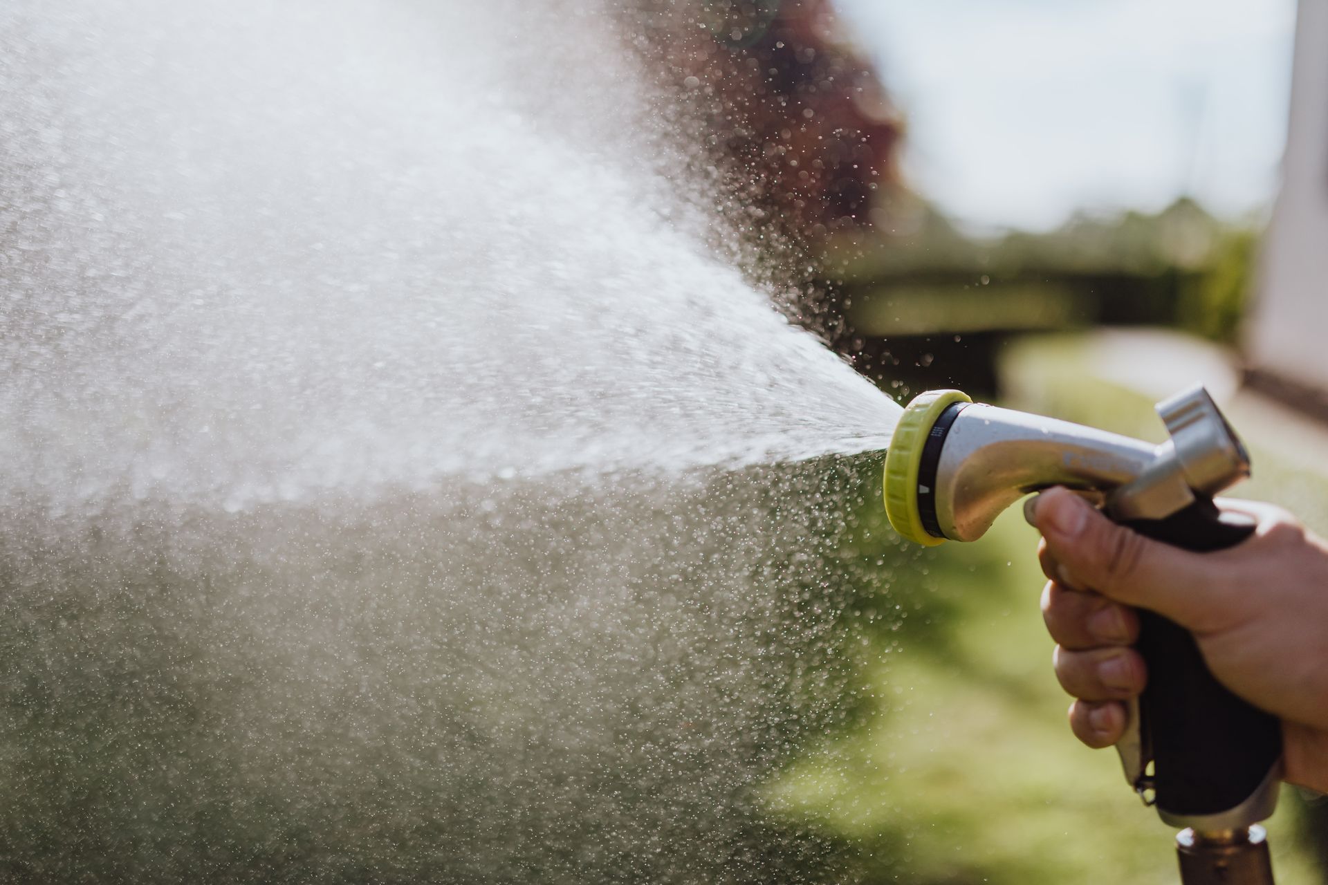 A hand holds a spray nozzle, misting water over a garden on a bright day.